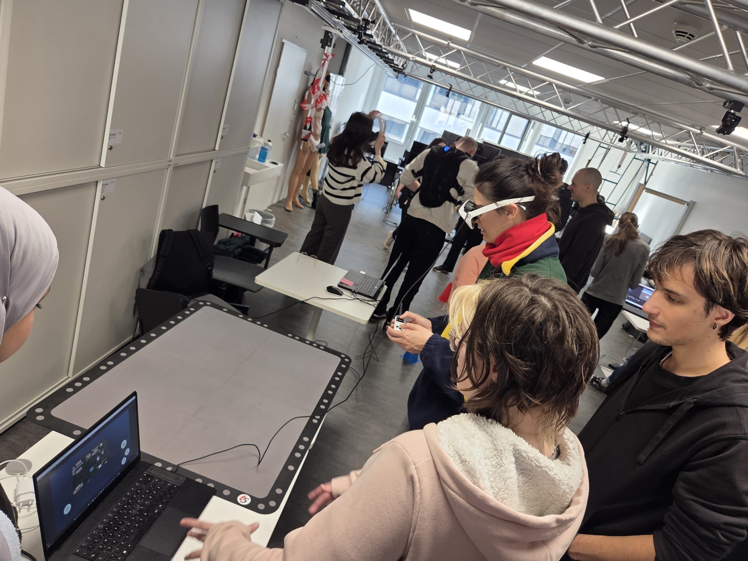 Eine Gruppe von Menschen interagiert mit Technologie und VR-Ausrüstung in einem modernen, überdachten Laborraum. Einige verwenden Laptops und VR-Headsets, während andere die Geräte beobachten oder bedienen. Der Raum ist hell beleuchtet und verfügt über verschiedene technische Einrichtungen.