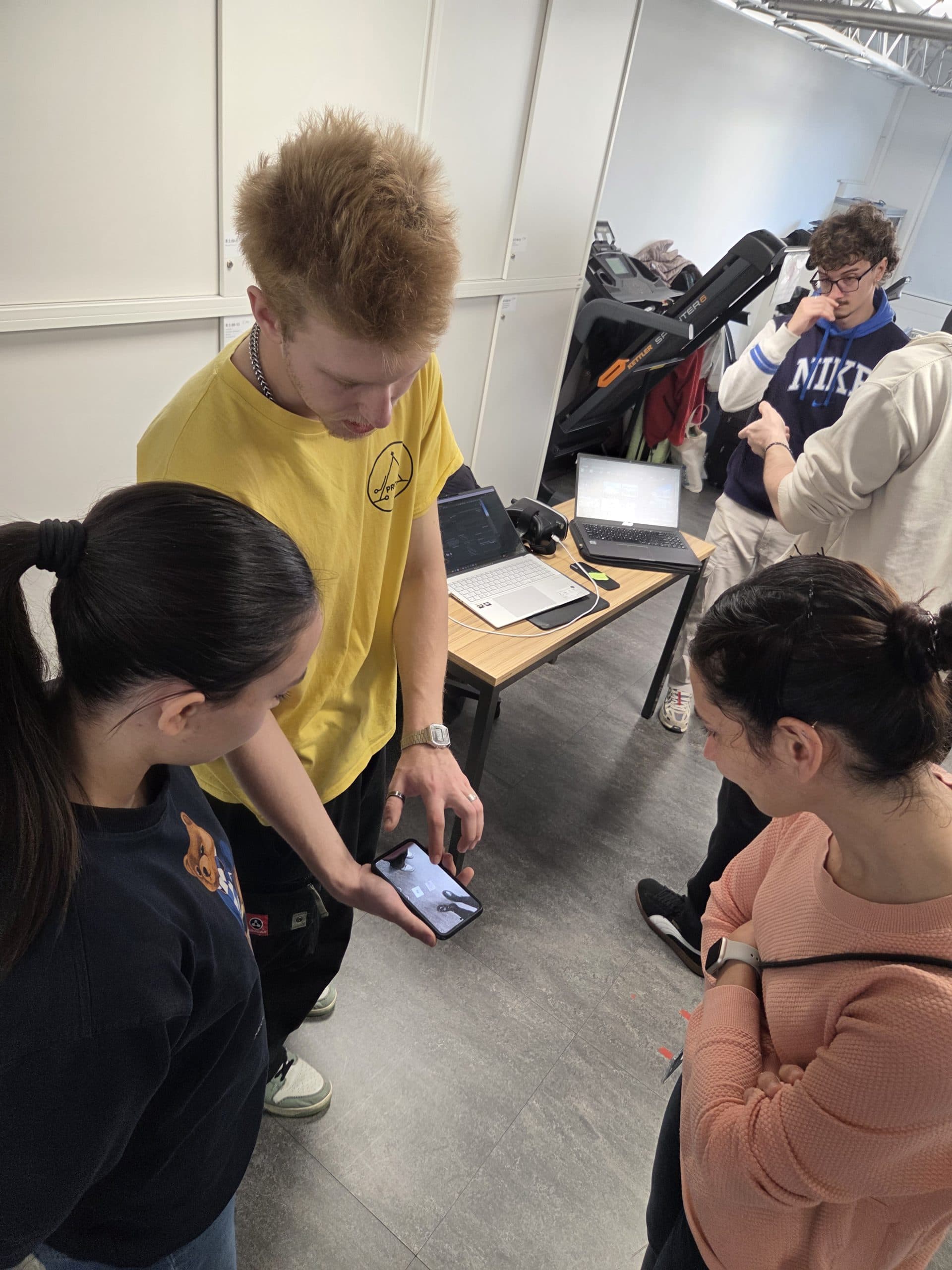 Drei Personen stehen zusammen und schauen auf ein Telefon, das von einem Mann in einem gelben Hemd gehalten wird, während sich zwei andere in der Nähe unterhalten. Hinter ihnen steht ein Tisch mit einem Laptop, einer Kamera, Kabeln und einem Rucksack.