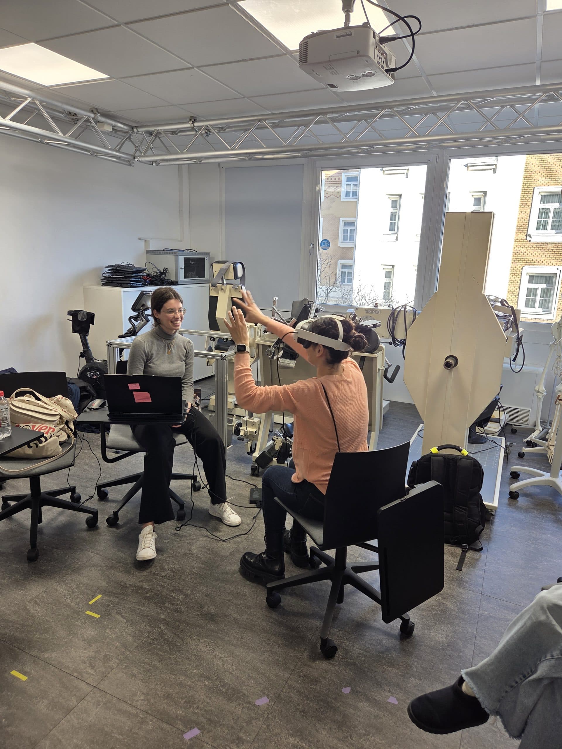 Zwei Menschen in einem hellen Labor, einer sitzt mit einem Laptop und lächelt, der andere sitzt und hält ein Gerät in die Höhe. Wissenschaftliche Geräte und Computer füllen den Raum, große Fenster zeigen Backsteinbauten draußen.