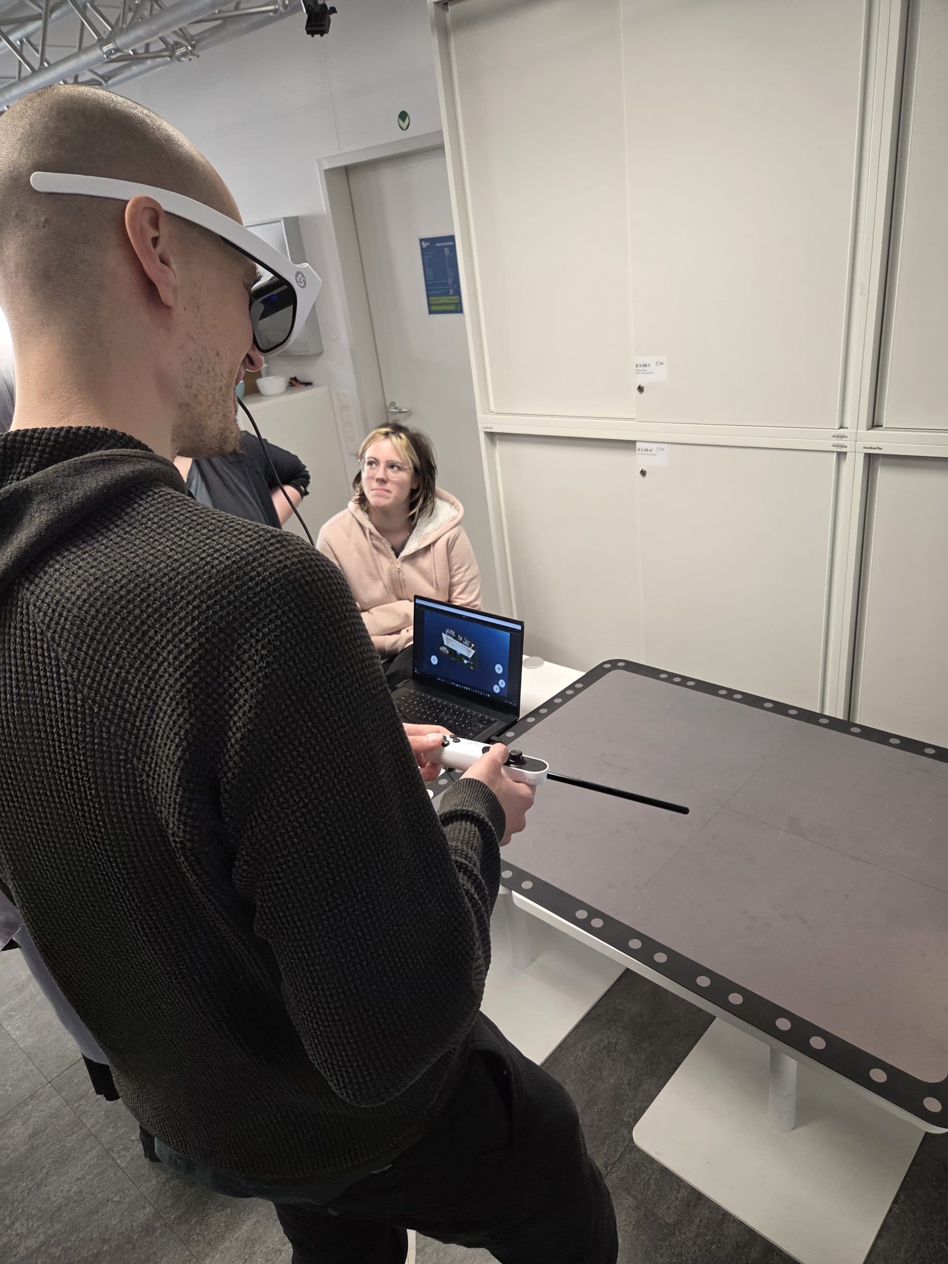 Eine Person, die eine AR-Brille trägt und einen Controller in der Hand hält, steht an einem Tisch mit einer digitalen Schnittstelle, während zwei Personen in der Nähe sitzen, von denen eine einen Laptop benutzt und zu der Person mit der Brille aufschaut.