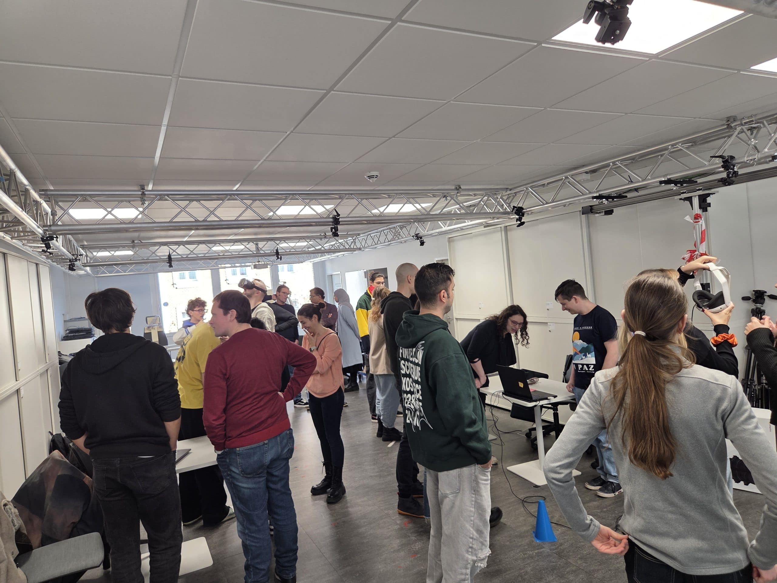 Eine Gruppe von Menschen steht in einem hellen, modernen Raum mit weißen Wänden und Decken und unterhält sich. Einige verwenden Laptops auf Tischen, während andere in kleinen Gruppen sprechen. Verschiedene Geräte und ein Stativ sind zu sehen.