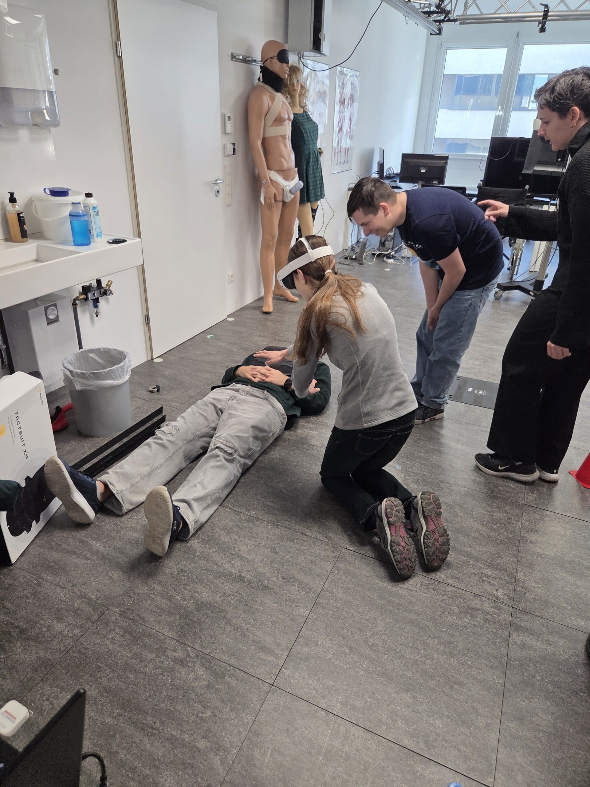 Eine Frau kniet neben einer am Boden liegenden Person und übt in einem medizinischen Schulungsraum die Wiederbelebung. Zwei weitere Personen beobachten, und im Hintergrund stehen medizinische Schaufensterpuppen.