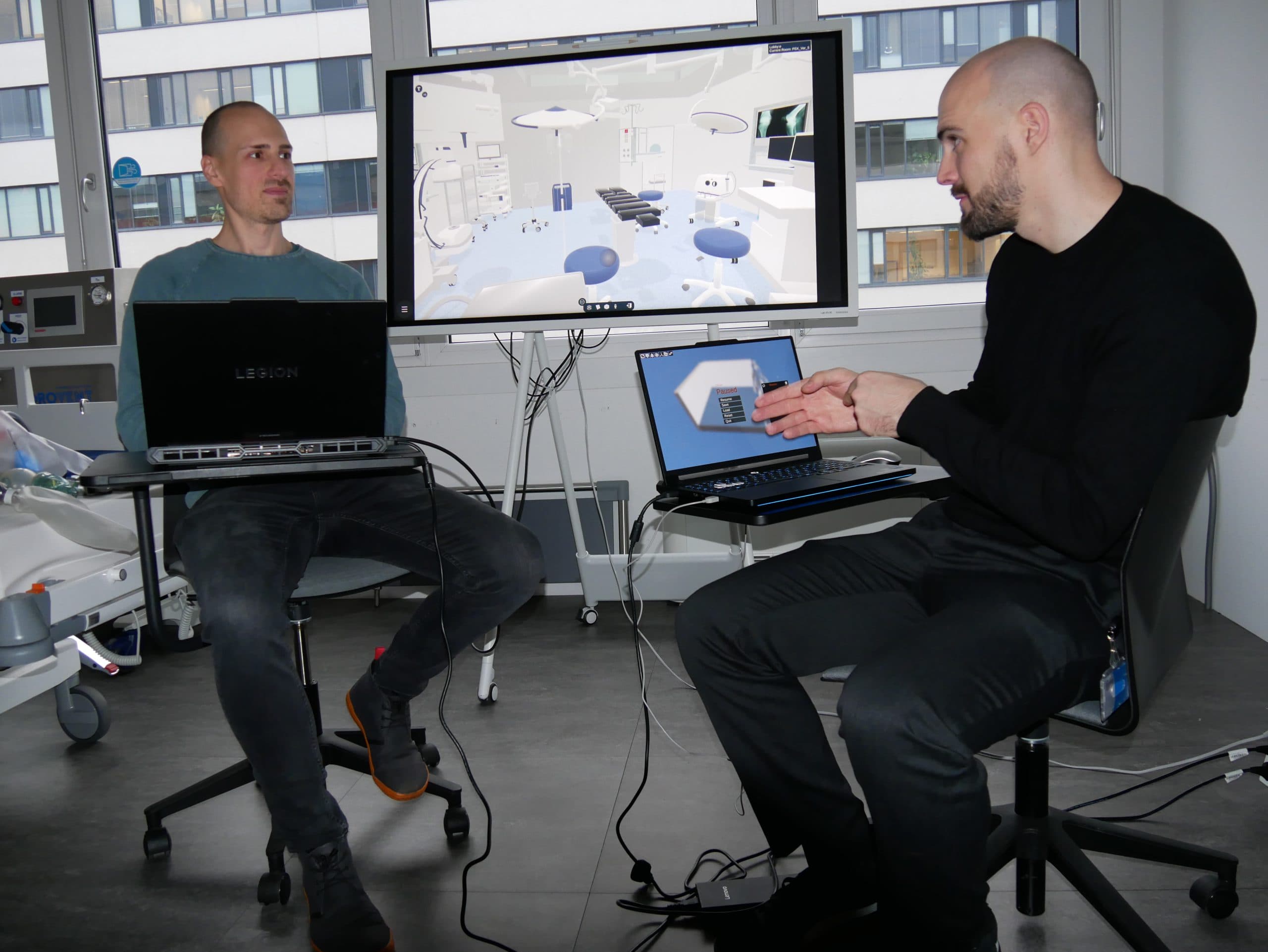 Zwei Männer sitzen sich mit Laptops in einem modernen Raum gegenüber. Hinter ihnen zeigt ein großer Monitor eine 3D-Darstellung einer medizinischen Einrichtung oder eines Operationssaals. Ein Mann gestikuliert mit seinen Händen, während sie diskutieren.