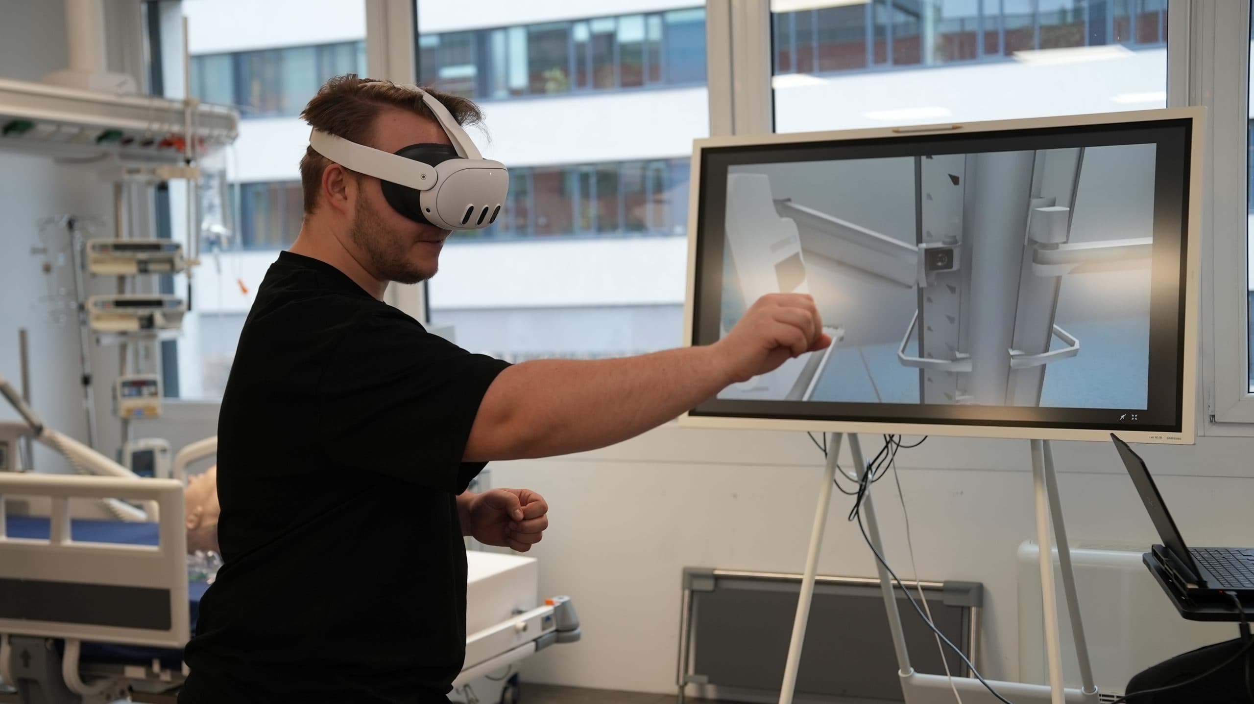 Eine Person, die ein Virtual-Reality-Headset trägt, interagiert mit einem Bildschirm, auf dem medizinische Geräte in einem modernen, gut beleuchteten Raum mit großen Fenstern angezeigt werden.