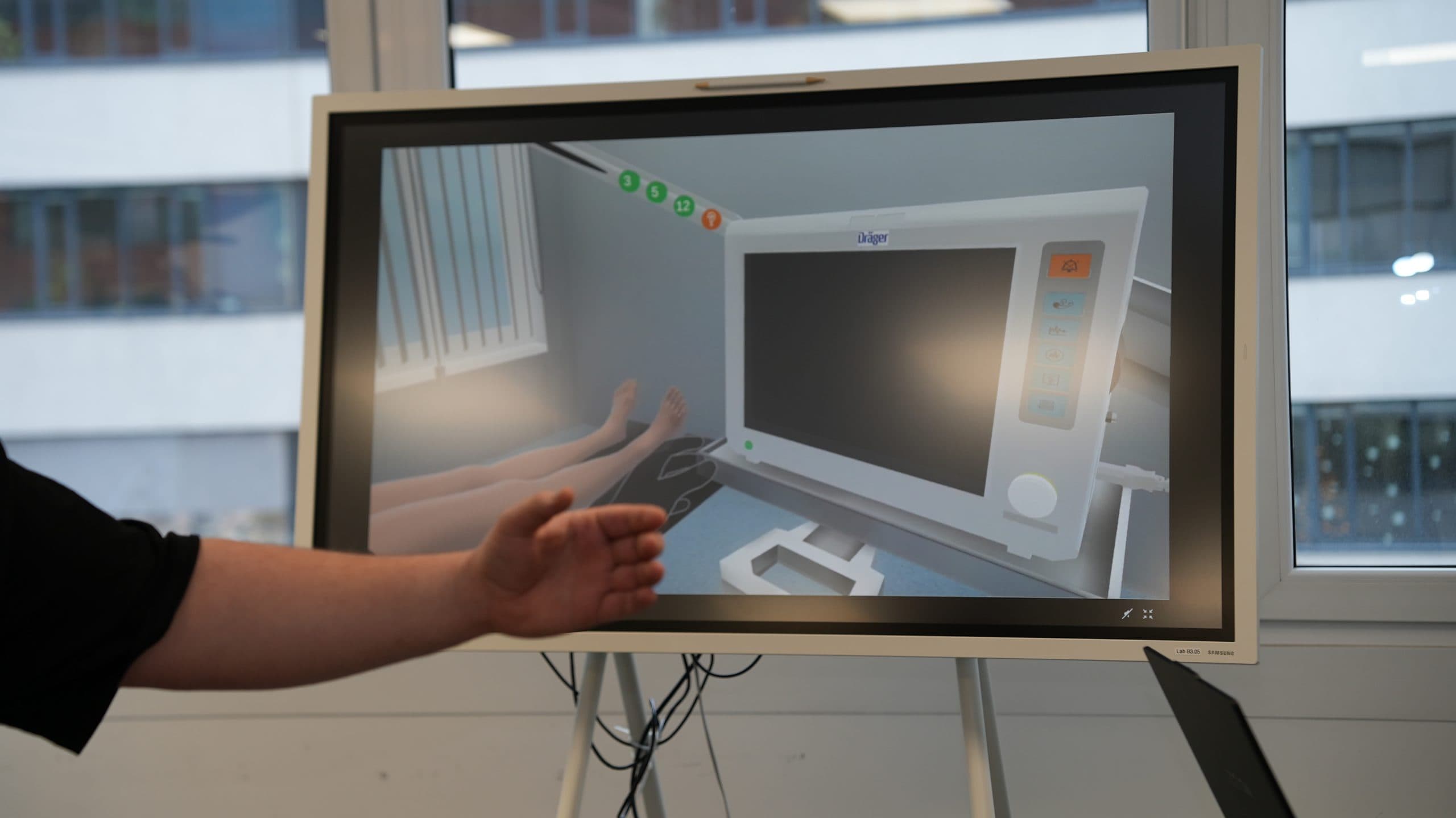 Eine Person gestikuliert in Richtung eines großen Bildschirms mit einer medizinischen Simulation, die einen Krankenhausmonitor und virtuelle Patientenbeine in einer klinischen Umgebung zeigt.