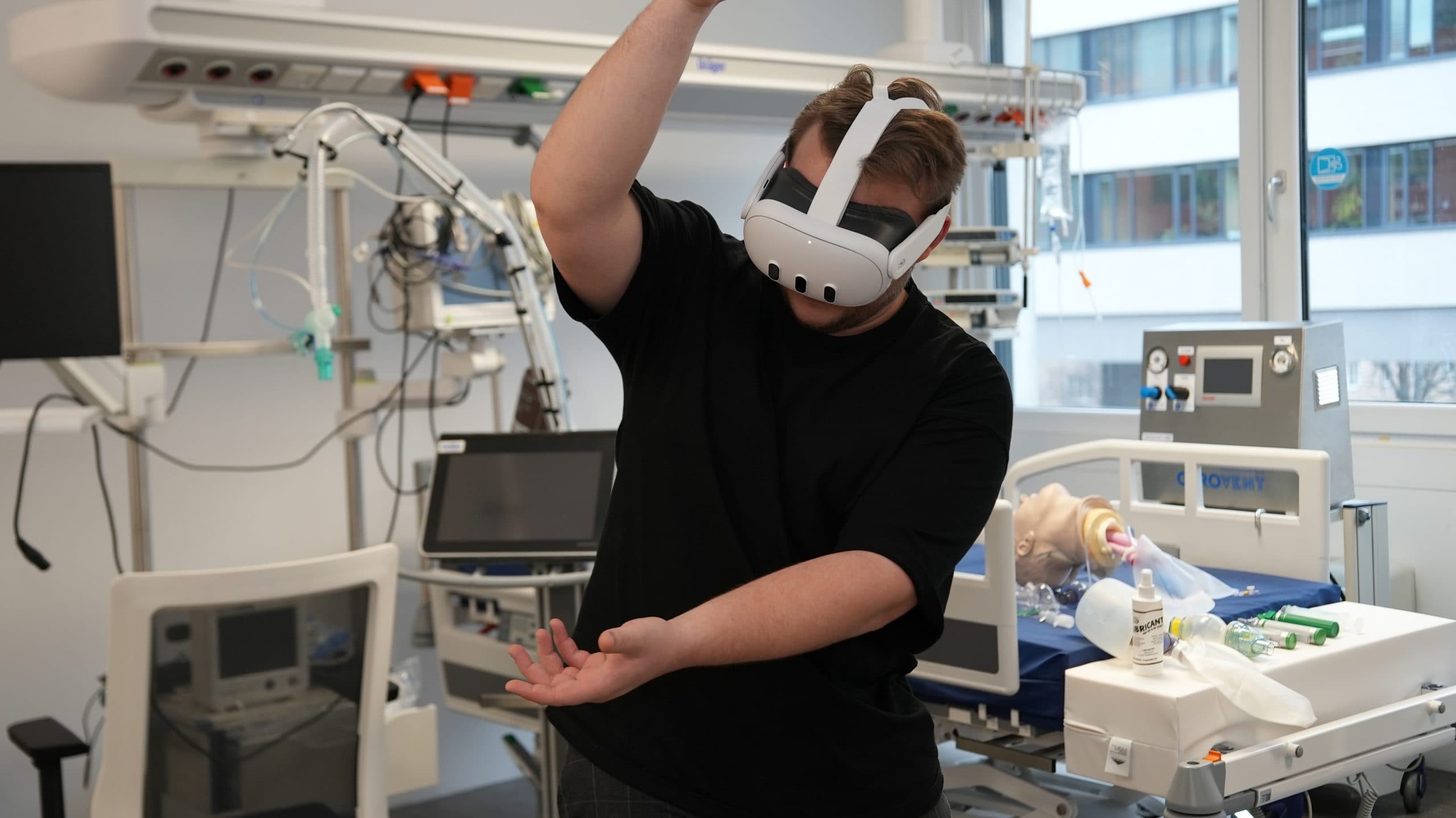Eine Person, die ein Virtual-Reality-Headset trägt, gestikuliert mit ihren Armen in einem Krankenhauszimmer mit medizinischen Geräten, darunter ein Krankenhausbett und eine medizinische Puppe.