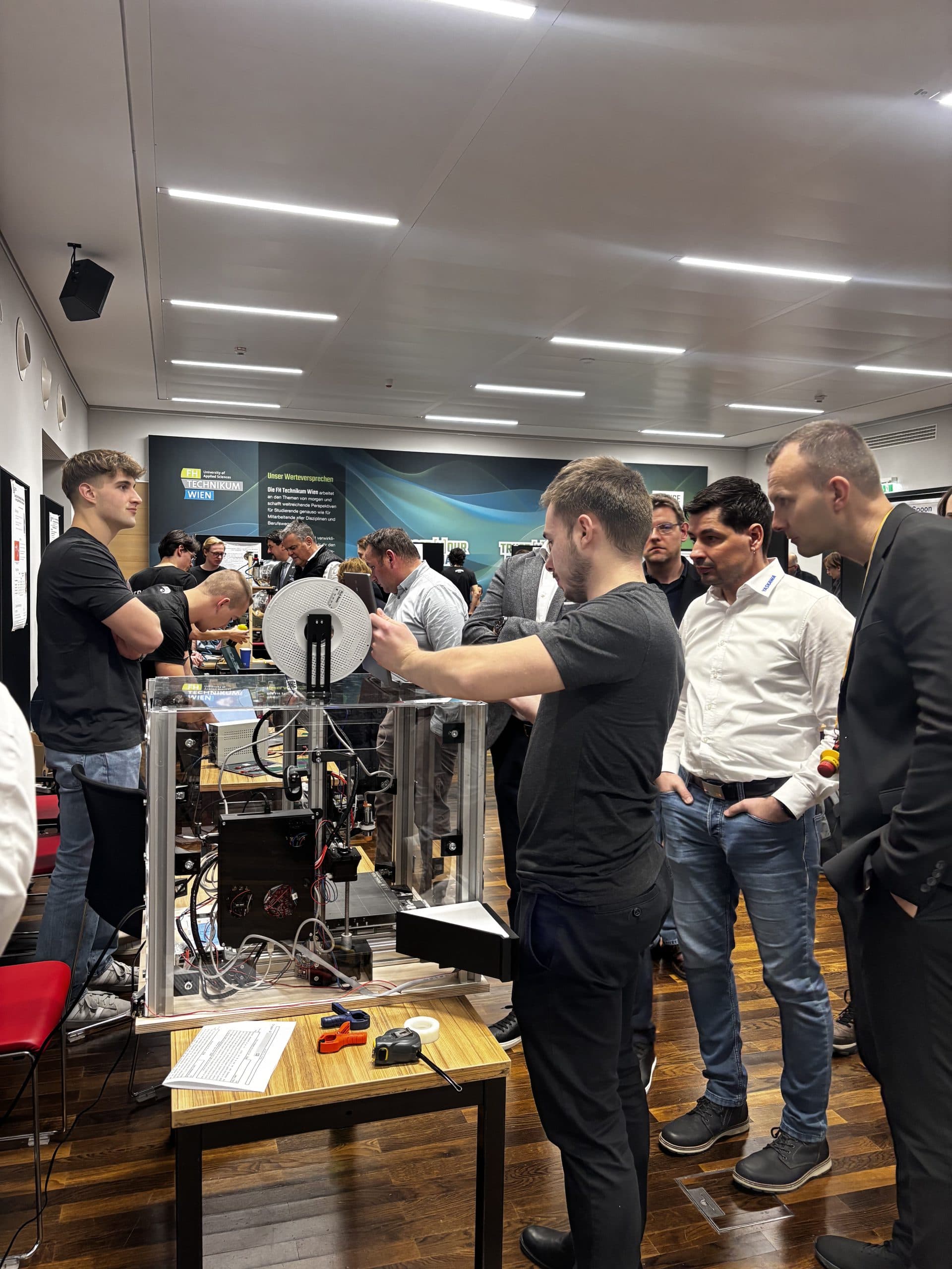Eine Gruppe von Menschen, meist Männer, steht auf einer Technologieausstellung um einen 3D-Drucker herum. Einige schauen genau zu, während eine Person das Gerät erklärt. Die Veranstaltung findet in einem Innenraum statt, mit Bannern und Displays im Hintergrund.