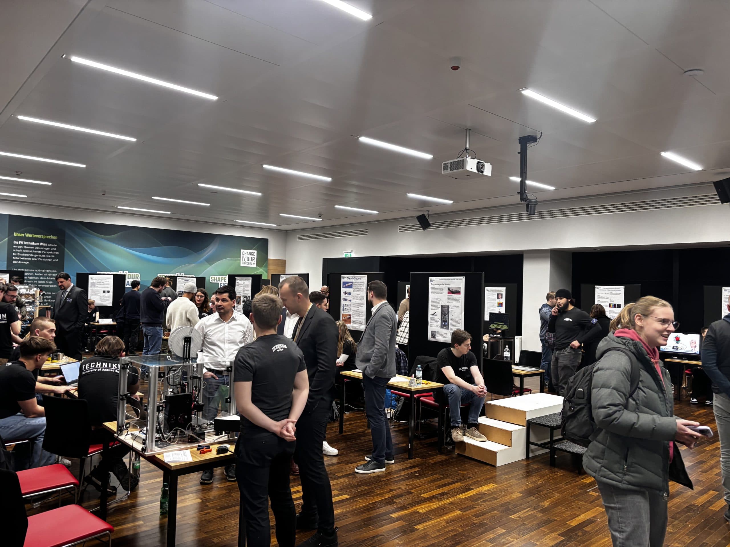 Eine geschäftige Veranstaltung in einer Halle, bei der Menschen stehen, sich unterhalten und verschiedene Technologieausstellungen und Poster betrachten. Einige Teilnehmer interagieren an Tischen mit Geräten, während andere durch den Raum gehen.