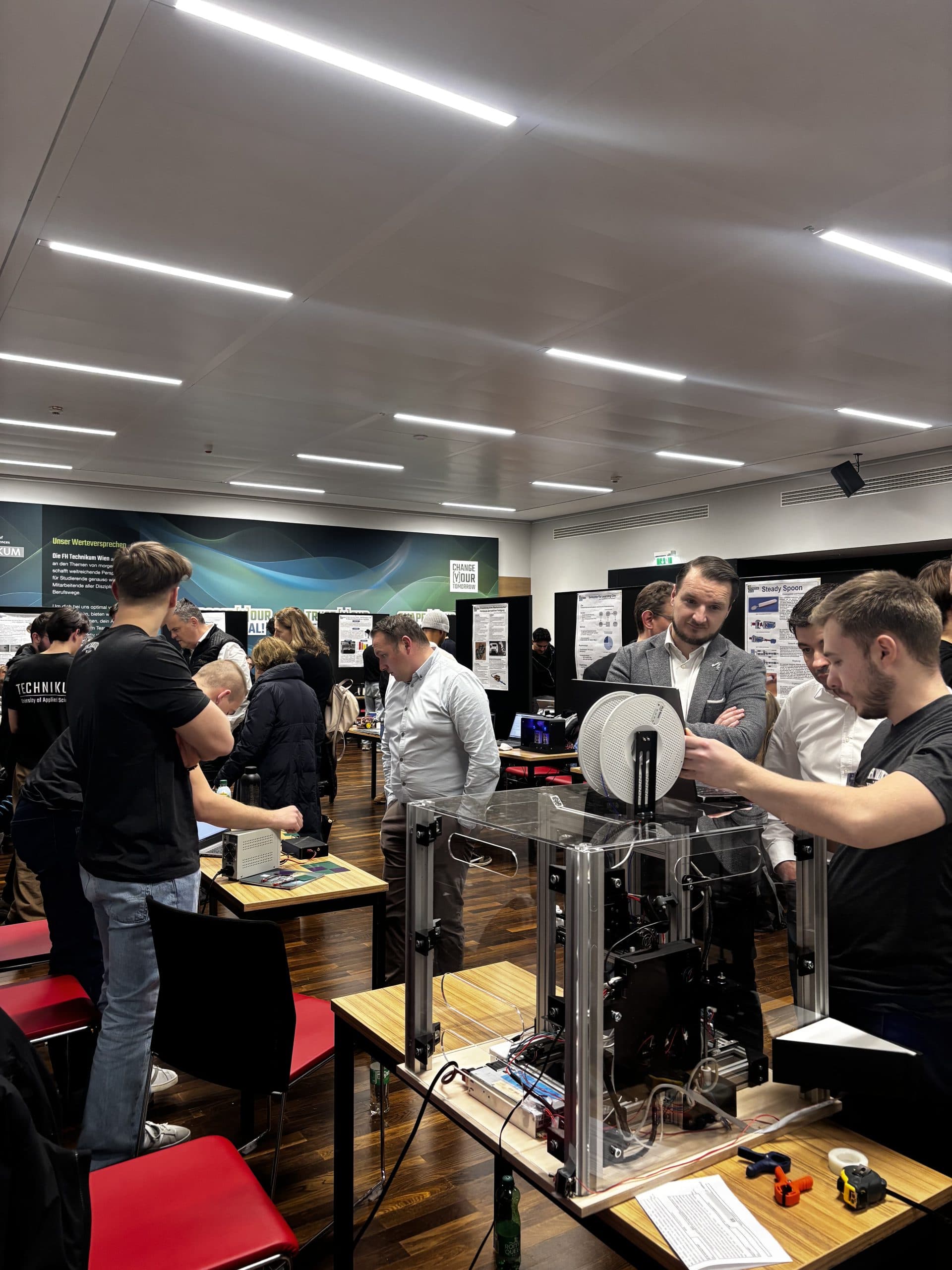 Auf einer Wissenschaftsmesse oder einer Ausstellung in einem hell erleuchteten Raum versammeln sich die Menschen um die Tische, an denen technische Projekte und 3D-Drucker ausgestellt sind, diskutieren miteinander und begutachten die Geräte und Poster.