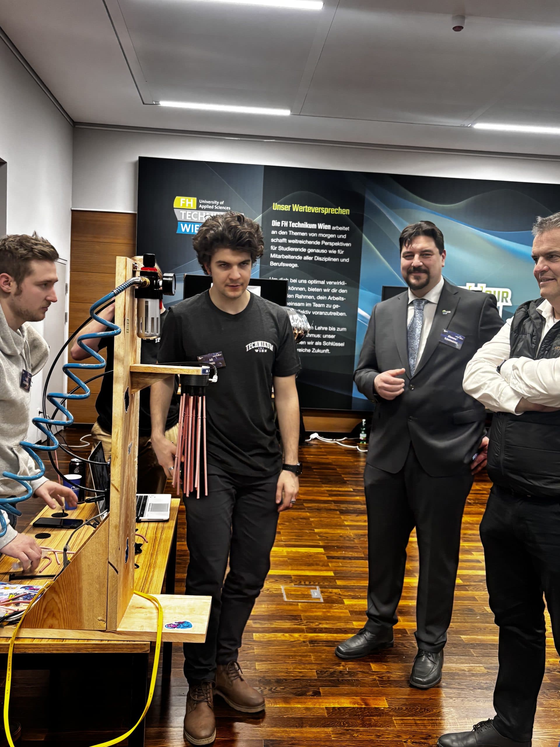 Vier Männer stehen um eine hölzerne Maschine mit Schläuchen und Drähten in einem modernen Raum. Ein Mann trägt einen Anzug, während die anderen leger gekleidet sind. Im Hintergrund sind ein Messestand und ein dunkler Holzboden zu sehen.