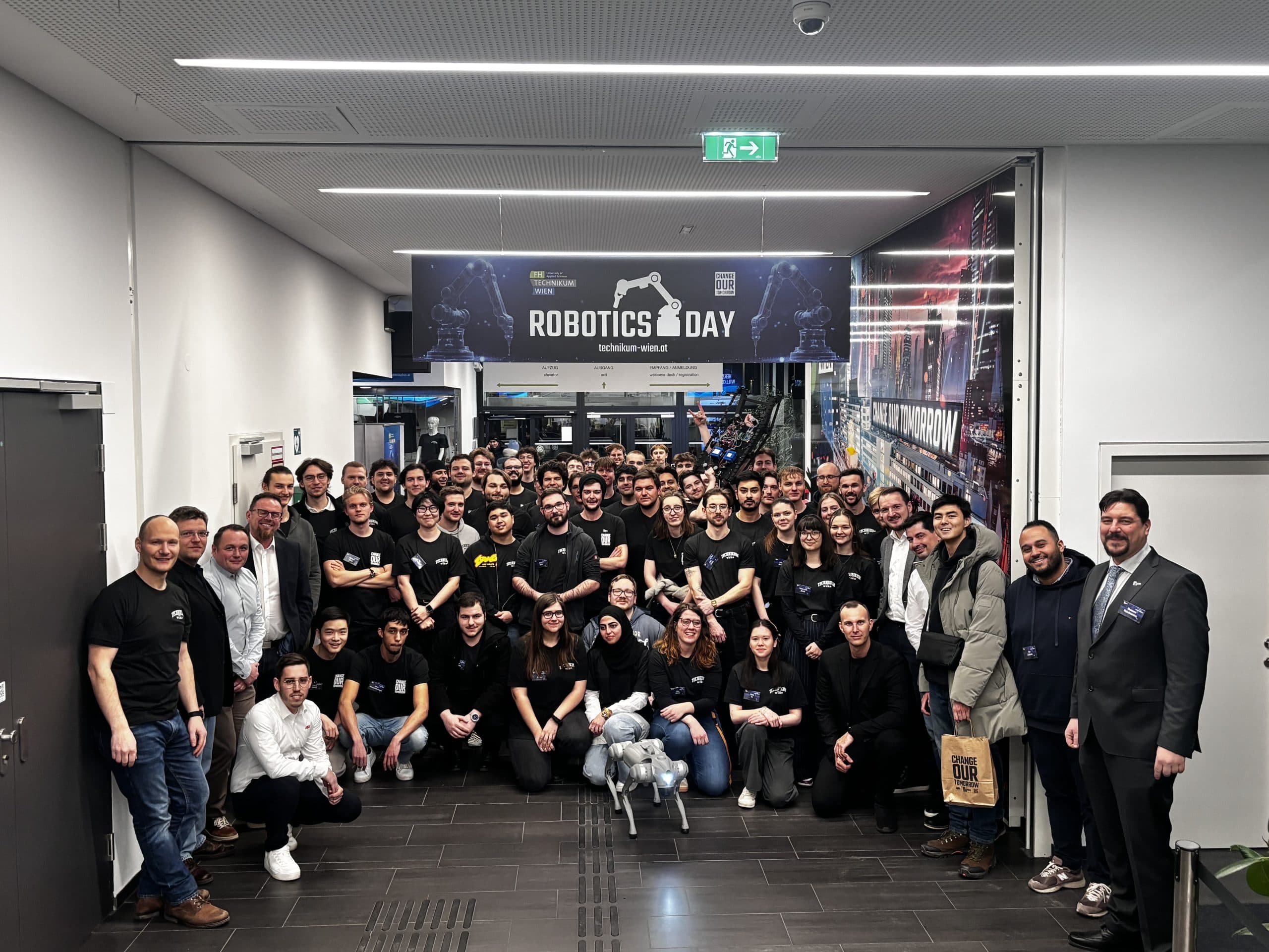 Eine große Gruppe von Menschen posiert in einem Flur unter einem Schild mit der Aufschrift 'Robotics Day'. Viele tragen schwarze T-Shirts, und vorne steht ein Roboterhund. Die Umgebung scheint eine moderne Indoor-Veranstaltung oder Konferenz zu sein.