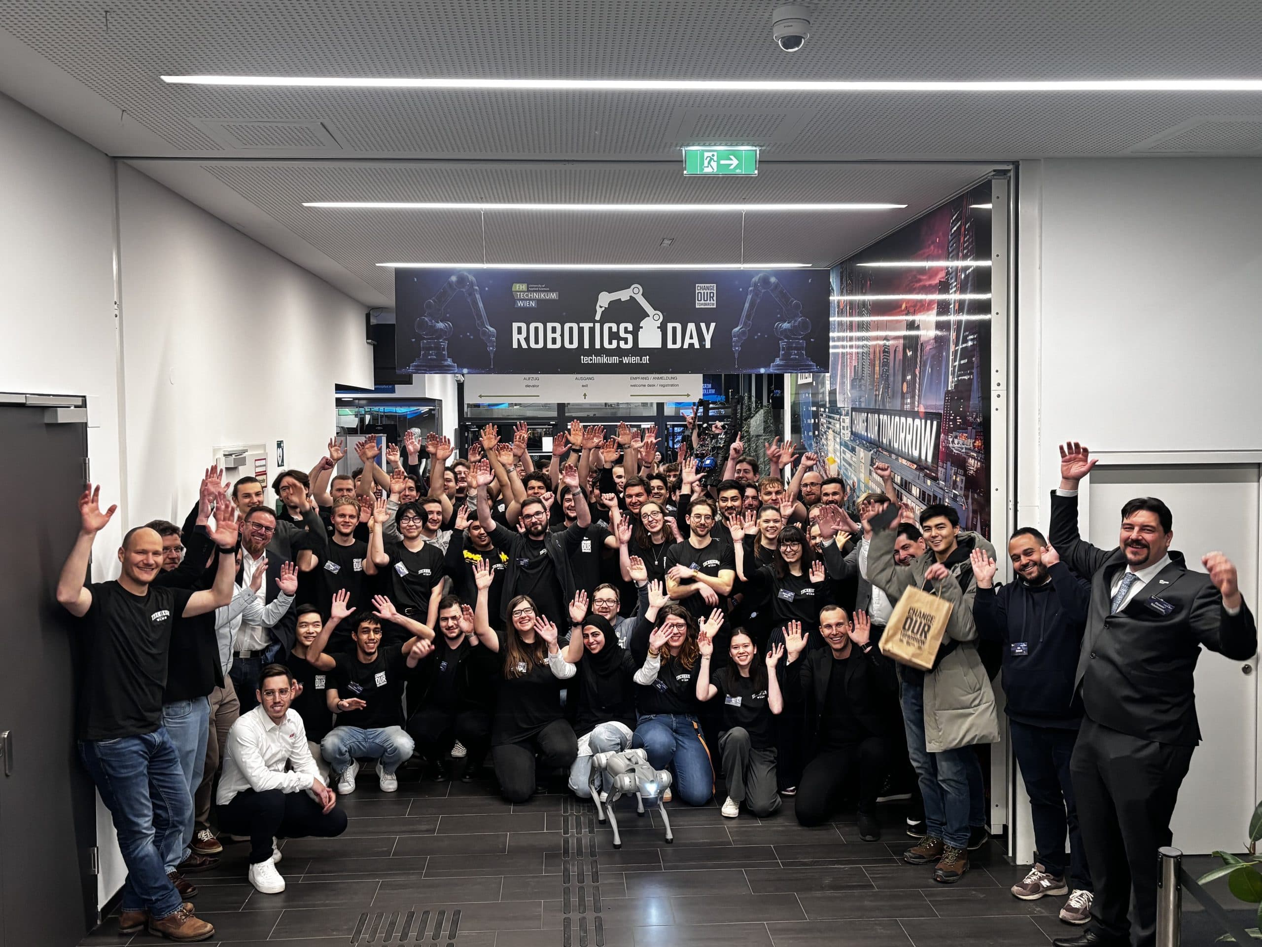 Eine große Gruppe von Menschen posiert unter einem Banner zum Tag der Robotik in einem Korridor, lächelt und hebt die Arme. Ein kleiner Roboterhund steht vor der Gruppe. Die Atmosphäre ist feierlich und energiegeladen.