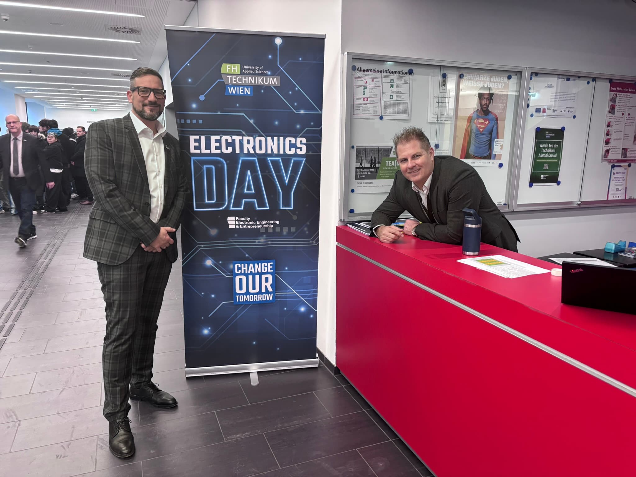 Zwei Männer in Anzügen stehen neben einem Banner mit der Aufschrift Electronics Day an einem roten Empfangstresen in einem modernen Flur. Auf dem Banner sind Logos und der Slogan 'Change Our Tomorrow' zu sehen. Im Hintergrund sind Menschen zu sehen.