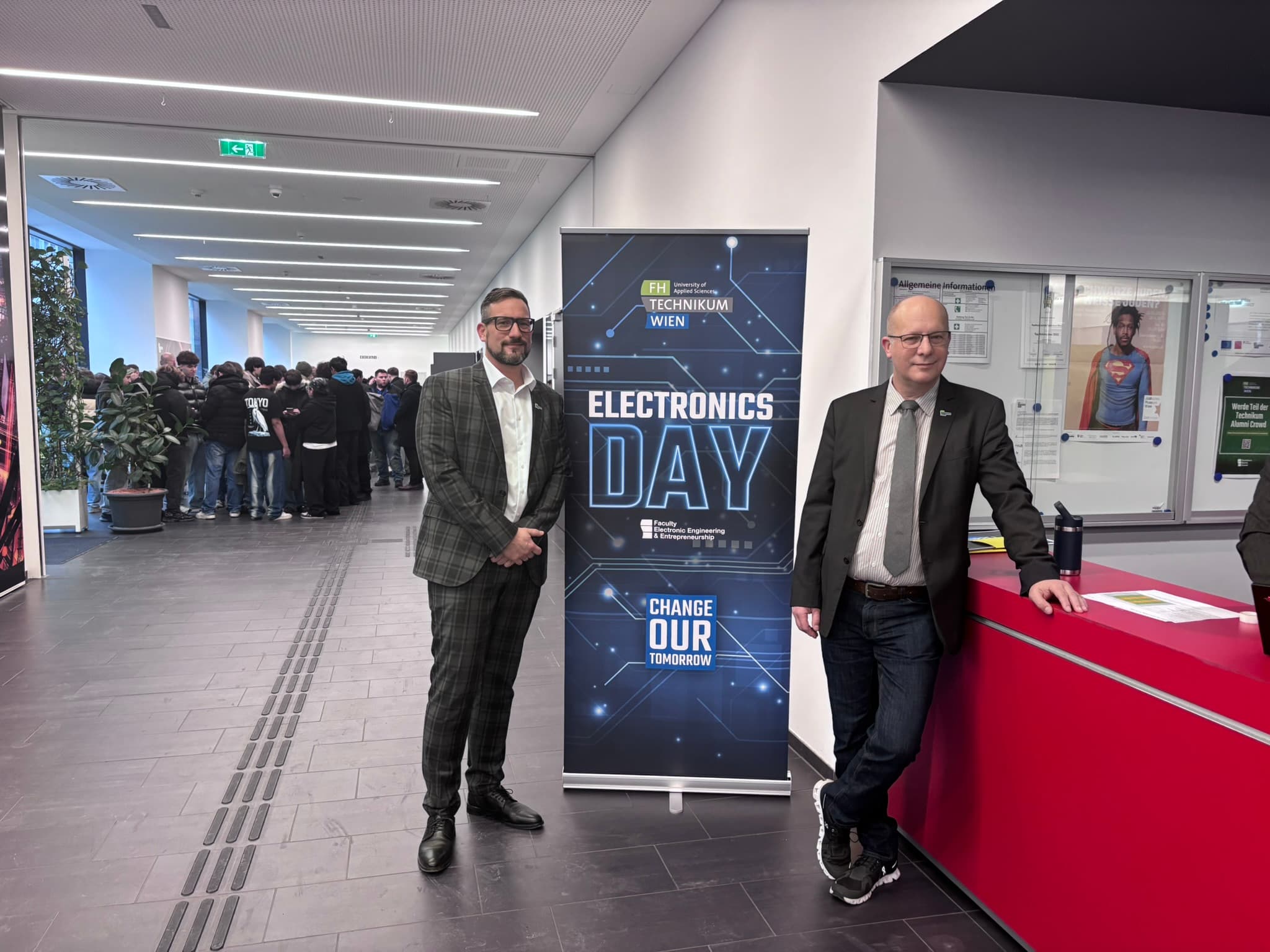 Zwei Männer in Geschäftskleidung stehen neben einem Schild mit der Aufschrift 'Electronics Day' in einem modernen Gebäude; einer steht neben einem roten Tresen, während sich im Hintergrund in der Nähe eines Flurs eine Gruppe von Menschen versammelt.