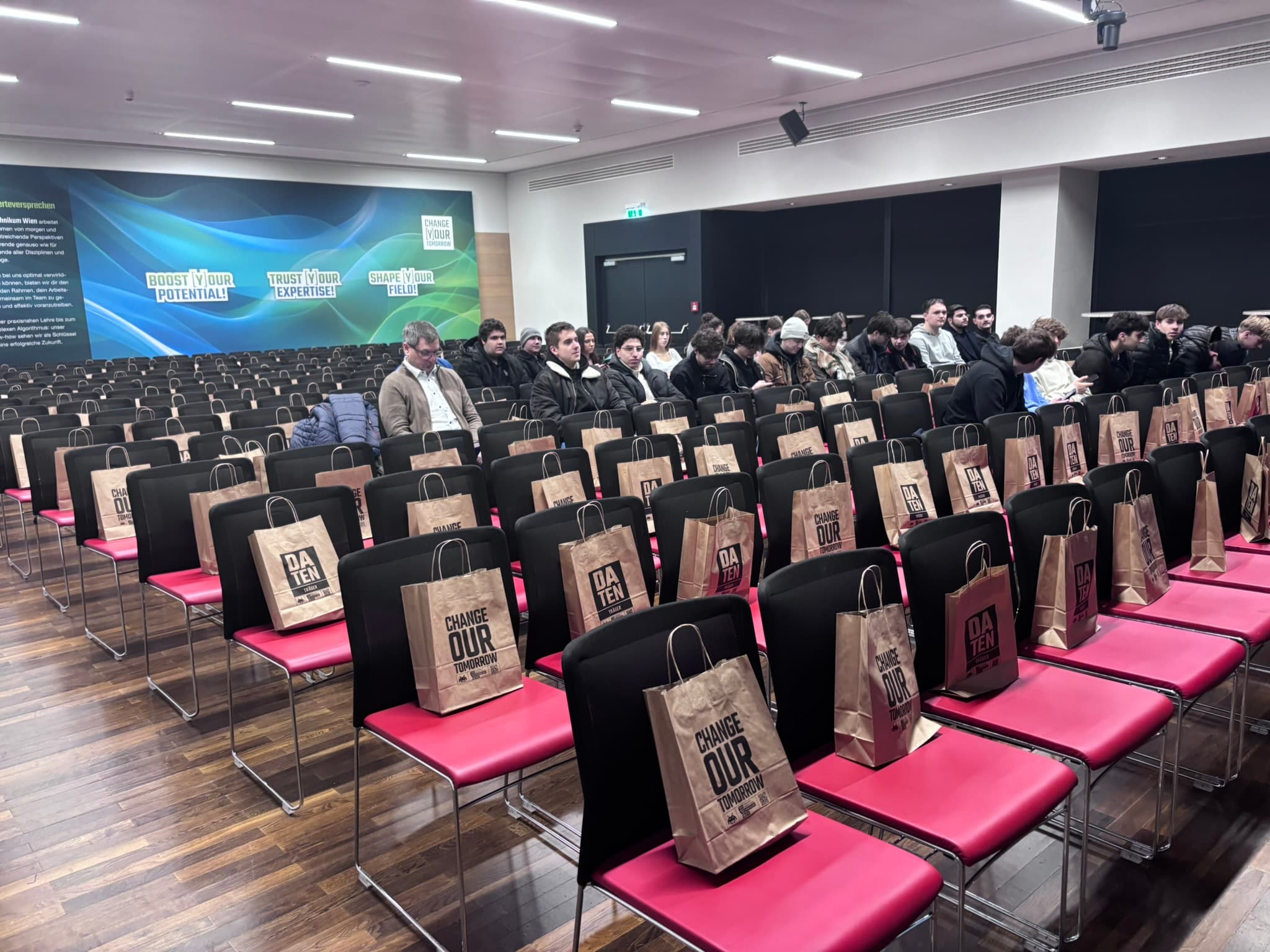 In einem Konferenzraum sitzt eine Gruppe von Menschen auf rosa und schwarzen Stuhlreihen. Auf vielen Sitzen liegen braune Papiertüten mit der Aufschrift Change Our. An der Rückwand hängt ein großes, buntes Banner.