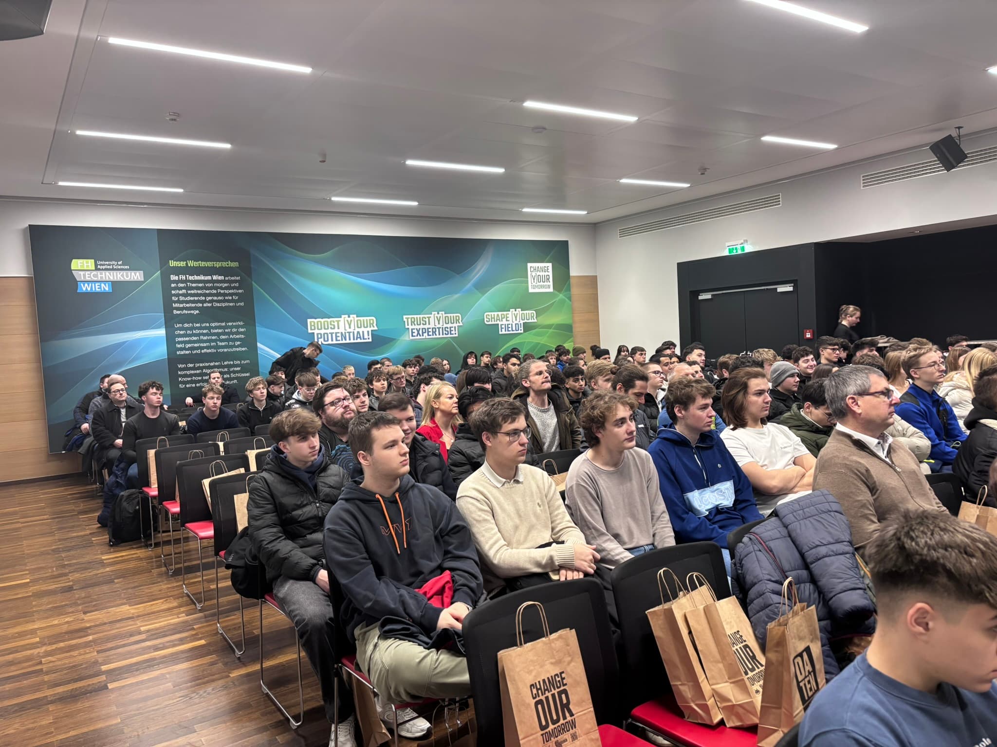 Eine große Gruppe von Menschen, meist junge Erwachsene, sitzt aufmerksam in Stuhlreihen bei einer Veranstaltung oder Präsentation in einem Gebäude. Auf einigen Sitzen liegen Geschenktüten, und die Wand im Hintergrund ist mit Motivationspostern geschmückt.