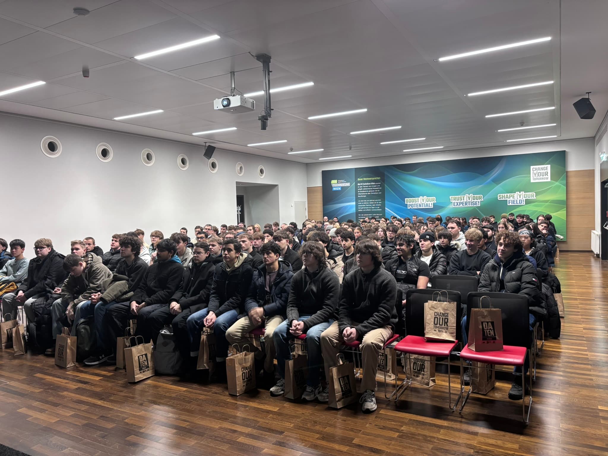 Eine große Gruppe von Menschen, meist junge Männer, sitzt in einem modernen Konferenzraum in Reihen mit Blickrichtung nach vorne, jeder mit einer Papiertüte auf dem Schoß; über ihnen ist ein Projektor an der Decke angebracht.