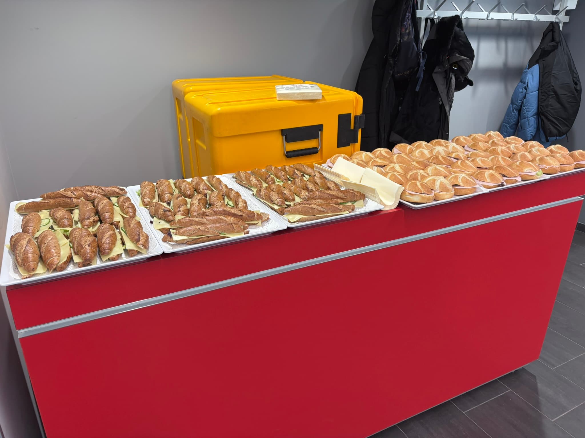 Auf einer roten Theke stehen Tabletts mit verschiedenen Sandwiches und Brötchen, dahinter steht ein großer gelber Lebensmittelcontainer. Mäntel hängen an Haken an der grauen Wand im Hintergrund.