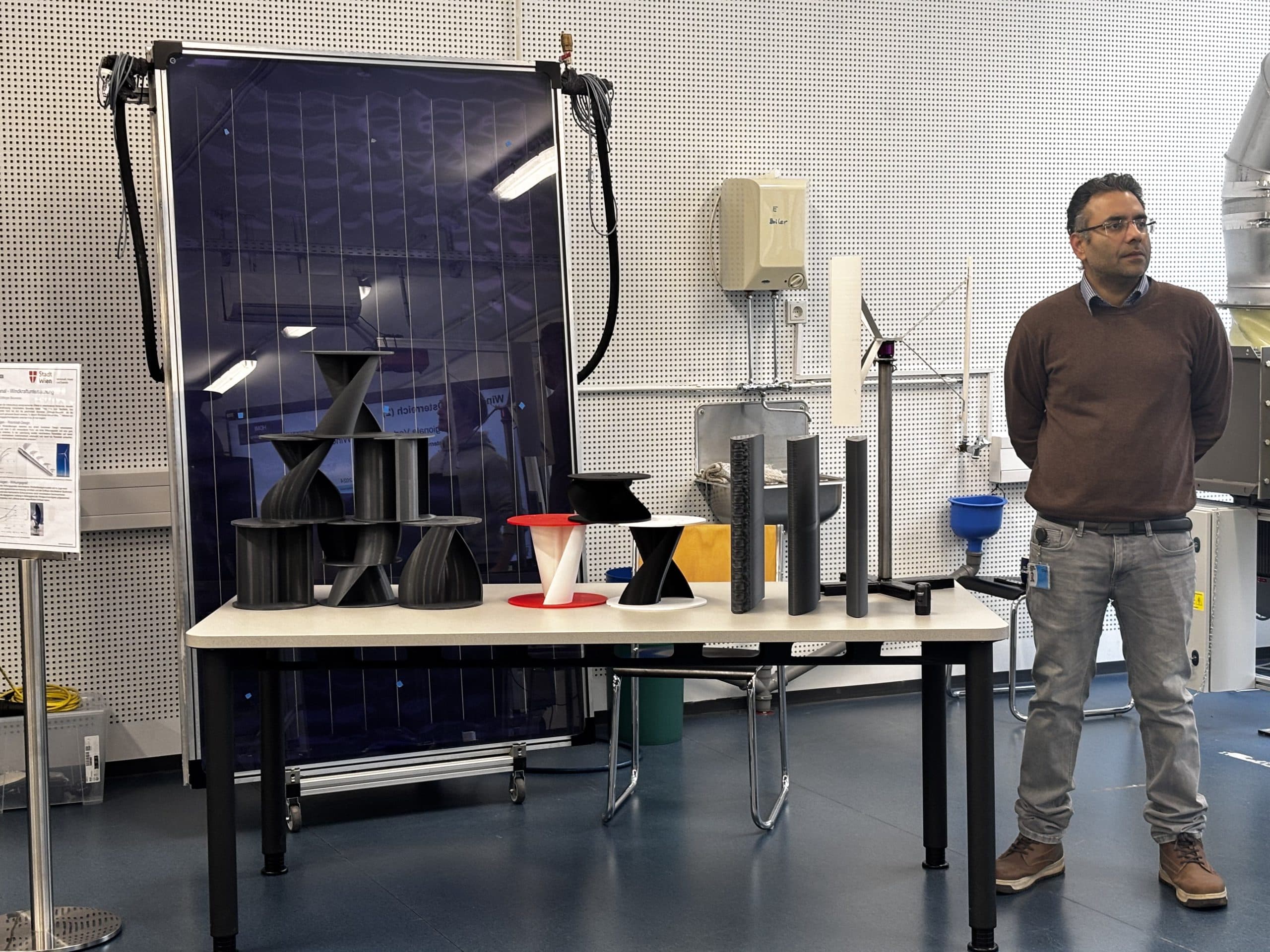 Ein Mann steht neben einem Tisch, auf dem verschiedene Modelle von Windturbinen-Prototypen in einem modernen Labor ausgestellt sind, mit einem großen Solarpanel und technischen Geräten im Hintergrund.