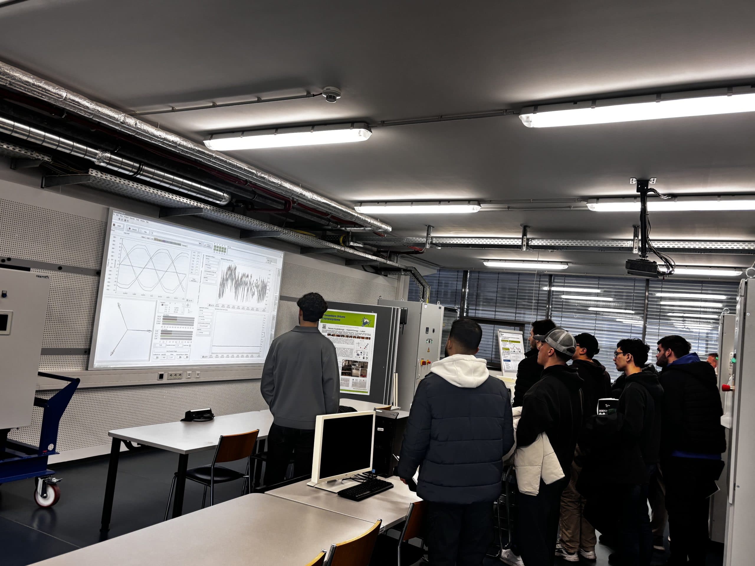 Eine Gruppe von Personen steht in einem modernen Klassenzimmer oder Labor und sieht sich eine Präsentation auf einem großen Bildschirm an, auf dem technische Diagramme und Daten angezeigt werden. In dem Raum sind auch Schreibtische, Computer und Poster zu sehen.