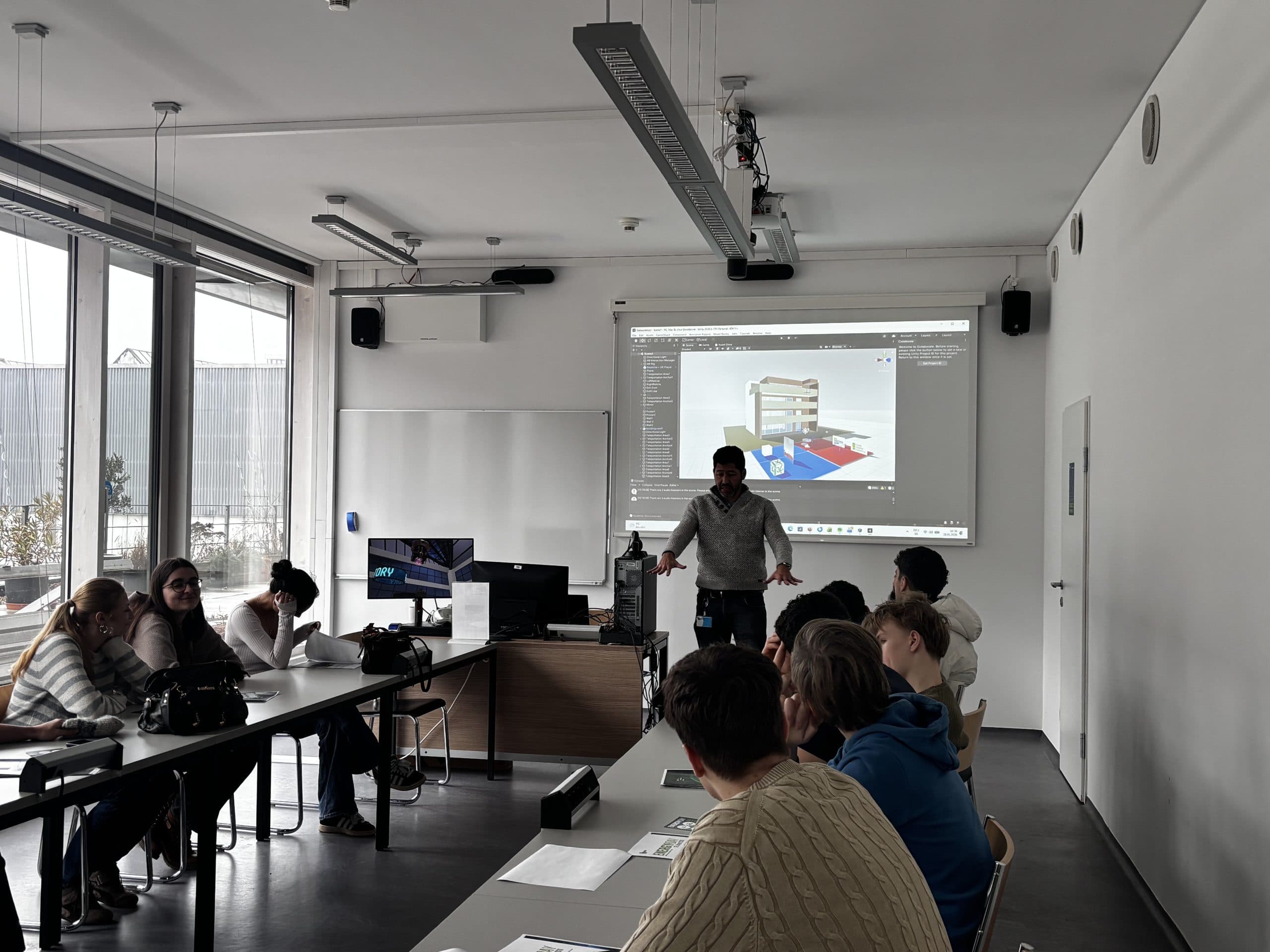 Ein Mann hält in einem modernen Klassenzimmer einen Vortrag vor einer Gruppe von Schülern. Ein 3D-Modell wird auf die Leinwand hinter ihm projiziert, und die Schüler sitzen an Tischen, hören zu und machen sich Notizen. Große Fenster lassen natürliches Licht herein.