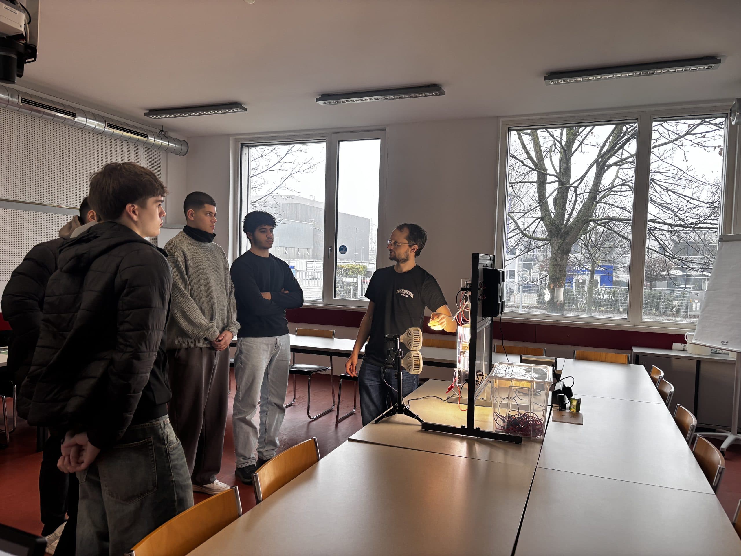 Vier Schüler stehen und hören einem Lehrer zu, der ein wissenschaftliches Experiment oder eine Demonstration mit Hilfe von Geräten auf einem Tisch im Klassenzimmer erklärt, wobei große Fenster und Bäume zu sehen sind.