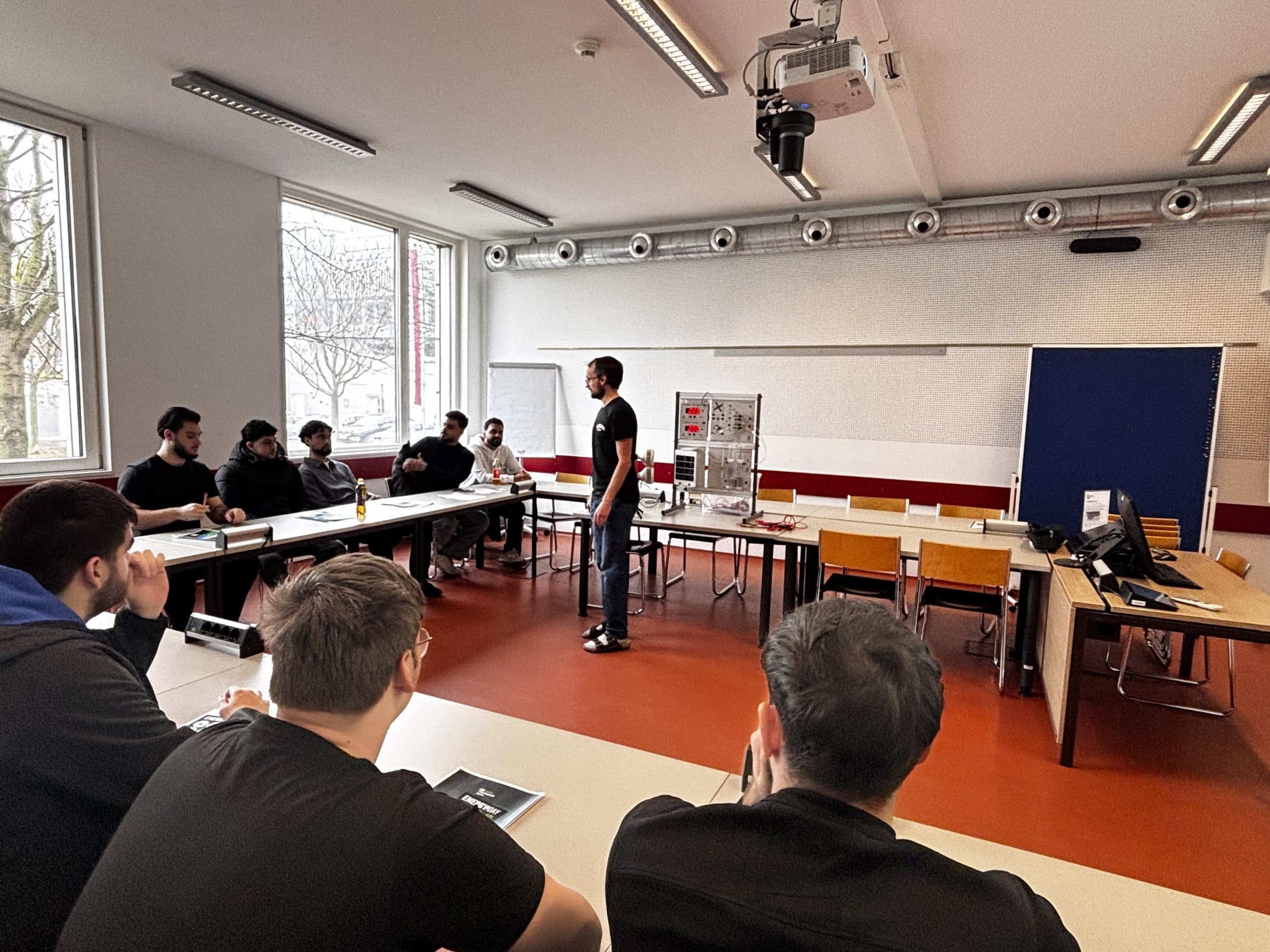 Eine Gruppe von Personen sitzt an Tischen in einem Klassenzimmer, während eine Person vor ihnen steht und spricht. In dem Raum befinden sich große Fenster, eine Tafel und Computer.