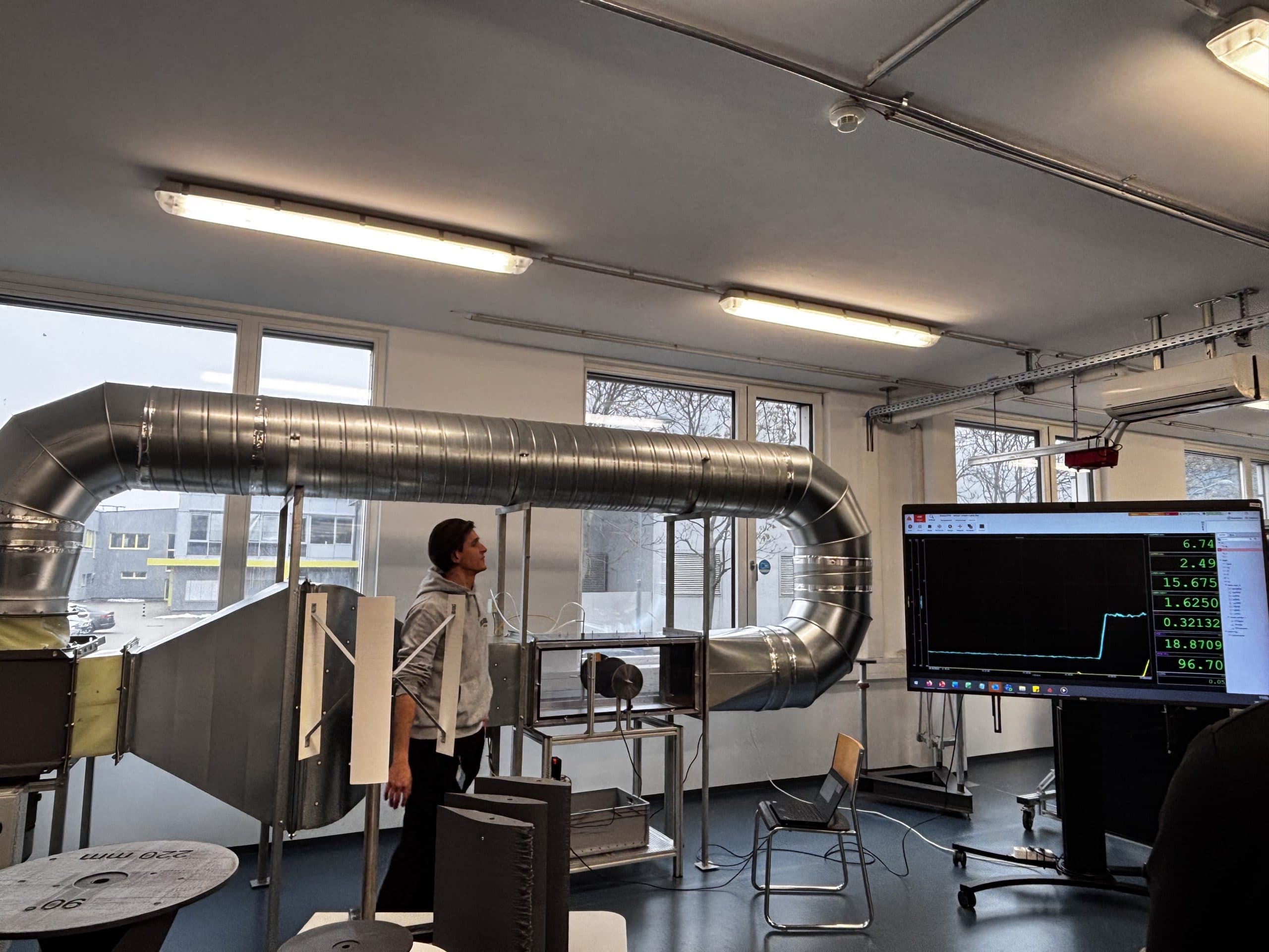 Eine Person steht in einem Labor mit großen Lüftungsschächten, Monitoren und Geräten. Auf einem Bildschirm werden Datendiagramme angezeigt. Im Hintergrund sind Fenster zu sehen, die eine bewölkte Außenszene zeigen.