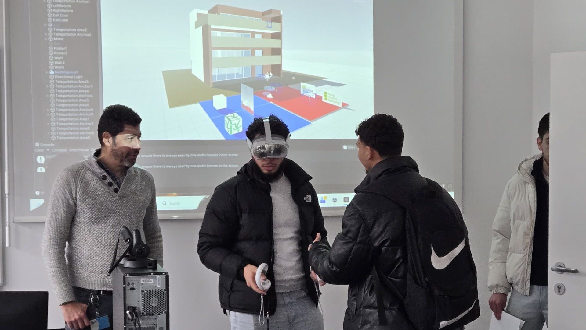 Vier Personen stehen in einem Klassenzimmer. Eine Person trägt ein VR-Headset, während die anderen zusehen. Ein Computerbildschirm hinter ihnen zeigt ein 3D-Gebäudemodell. Auf dem Tisch davor ist eine Ausrüstung aufgebaut.