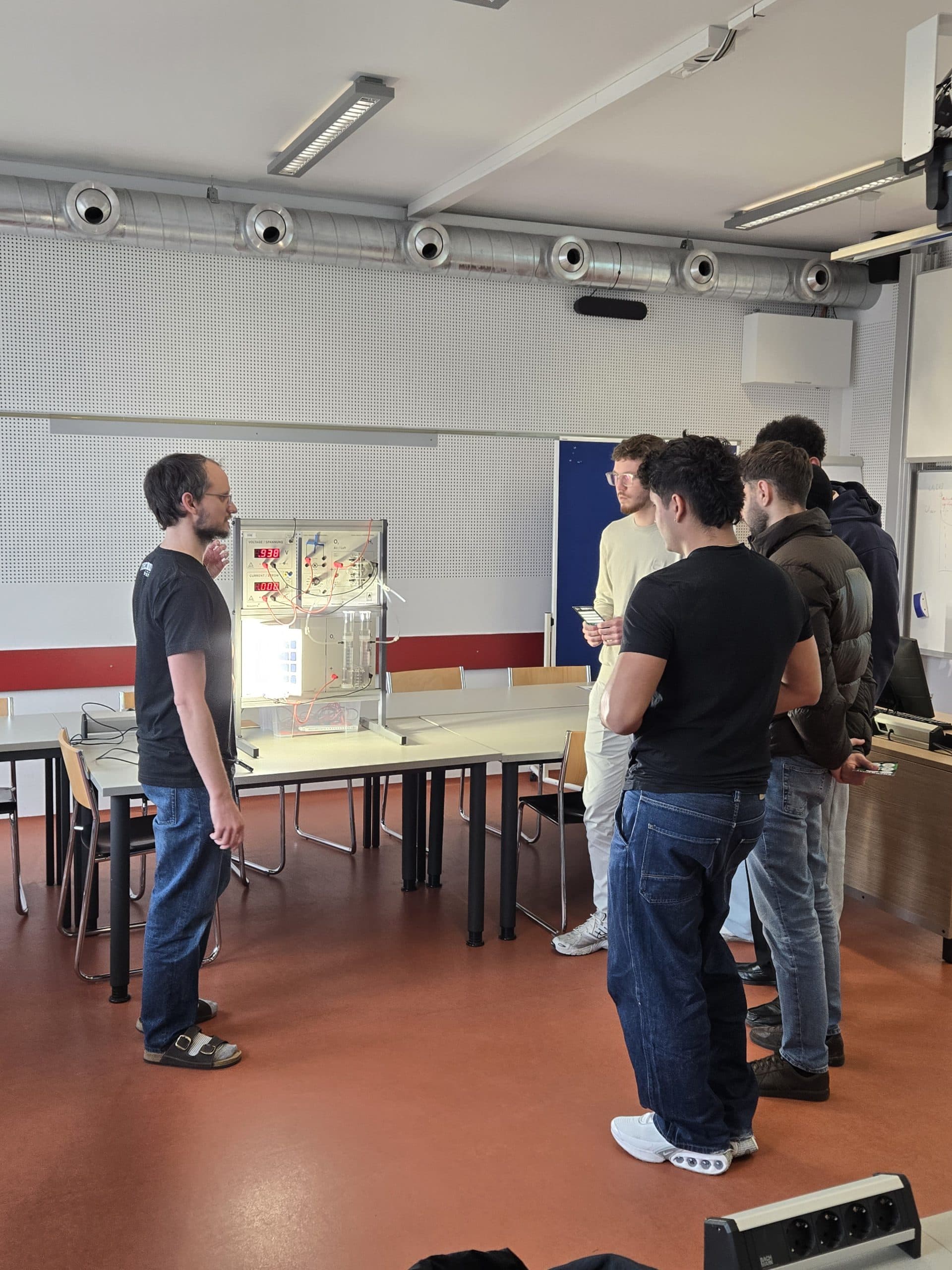 Eine Gruppe von fünf Personen steht in einem Klassenzimmer um eine Glasvitrine auf einem Tisch und führt eine Diskussion. Eine Person wendet sich an die Gruppe, während die anderen aufmerksam zuhören.