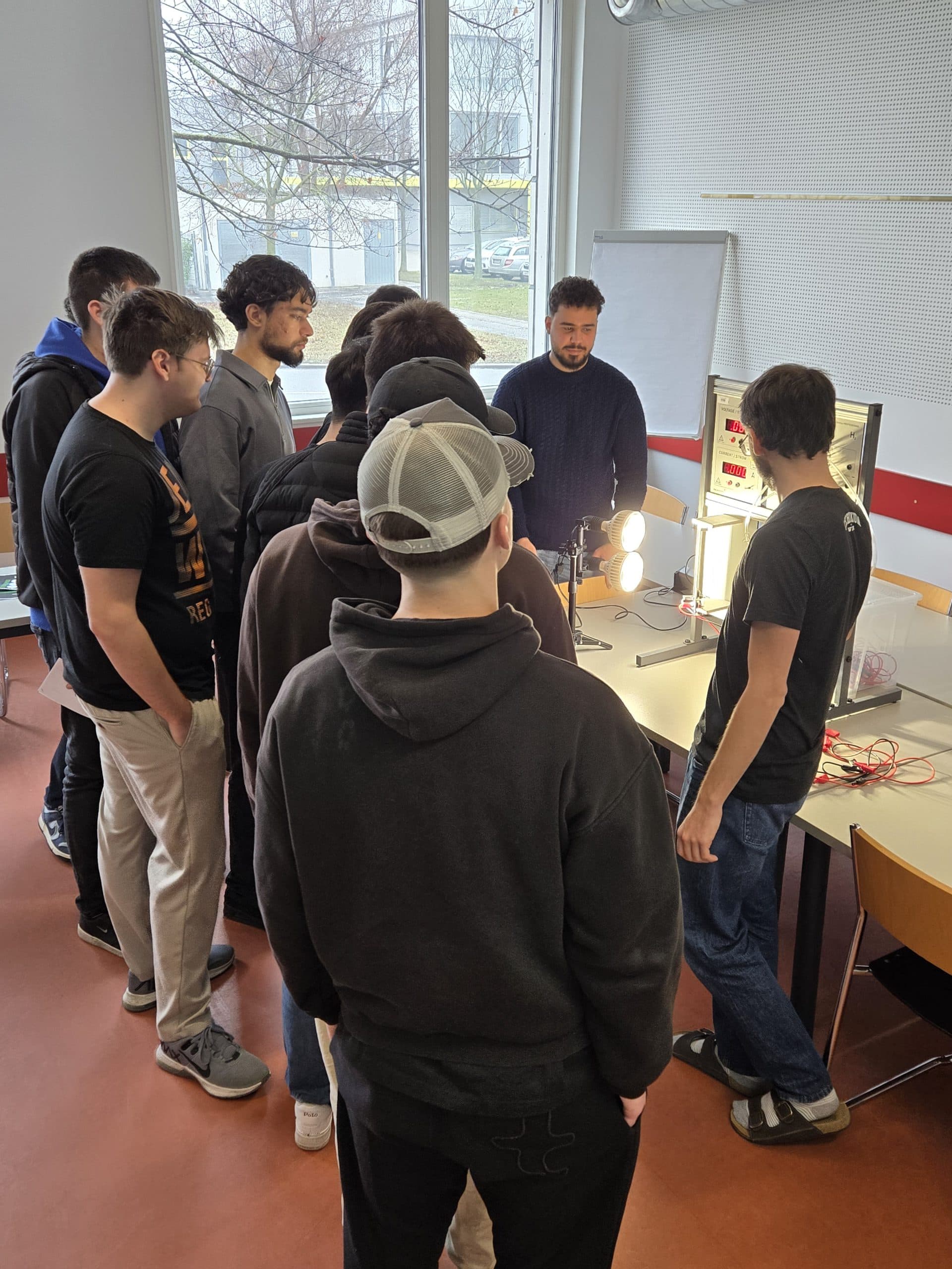 Eine Gruppe von Schülern steht um einen Tisch herum und beobachtet aufmerksam einen Mann, der in einem Klassenzimmer mit großen Fenstern ein elektrisches Experiment oder Gerät vorführt.
