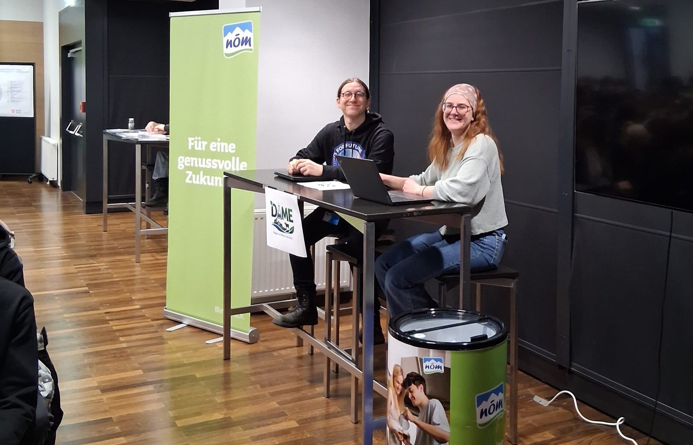 Zwei Frauen sitzen an einem hohen Tisch auf einer Bühne und lächeln dem Publikum zu. Auf einem grünen Transparent neben ihnen steht Für eine genussvolle Zukunft. Es scheint sich um eine Präsentation oder eine Podiumsdiskussion zu handeln.
