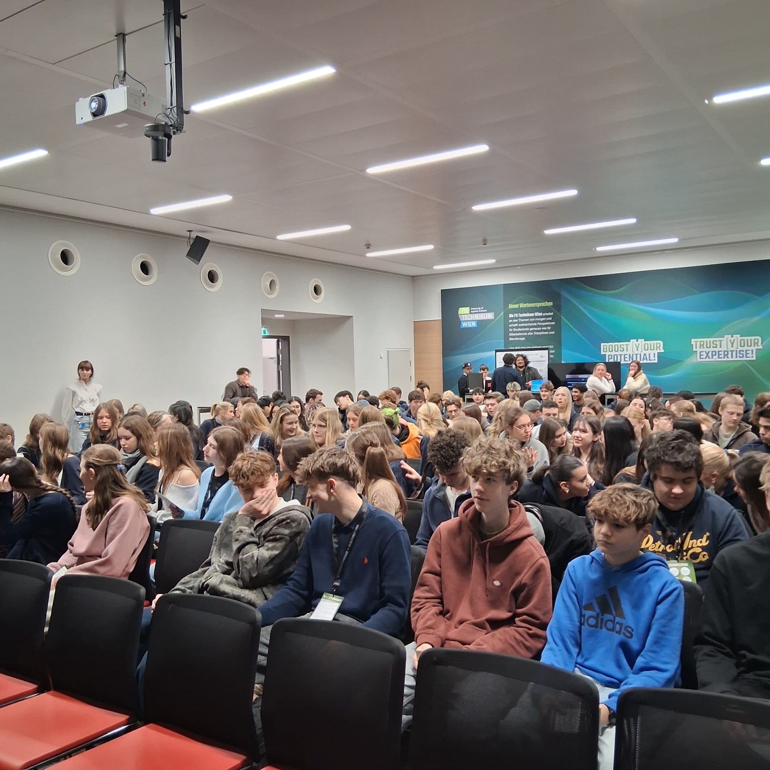 Eine große Gruppe junger Menschen sitzt in Stuhlreihen in einem modernen Konferenzraum und blickt nach vorne. Ein paar Erwachsene stehen hinten an einer Wand mit Schildern, auf denen 'Trust Your Expertise' steht.