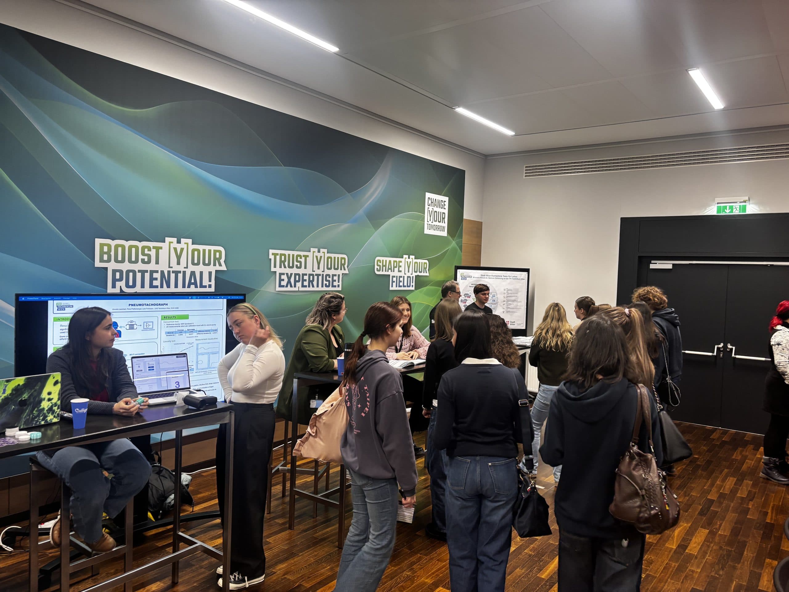 Eine Gruppe von Menschen versammelt sich in einem modernen Raum mit Holzfußboden, unterhält sich und knüpft Kontakte an Stehtischen. An der Wand hängen Schilder mit den Aufschriften 'BOOST YOUR POTENTIAL!', 'TRUST YOUR EXPERTISE!' und 'SHAPE YOUR FIELD!'.