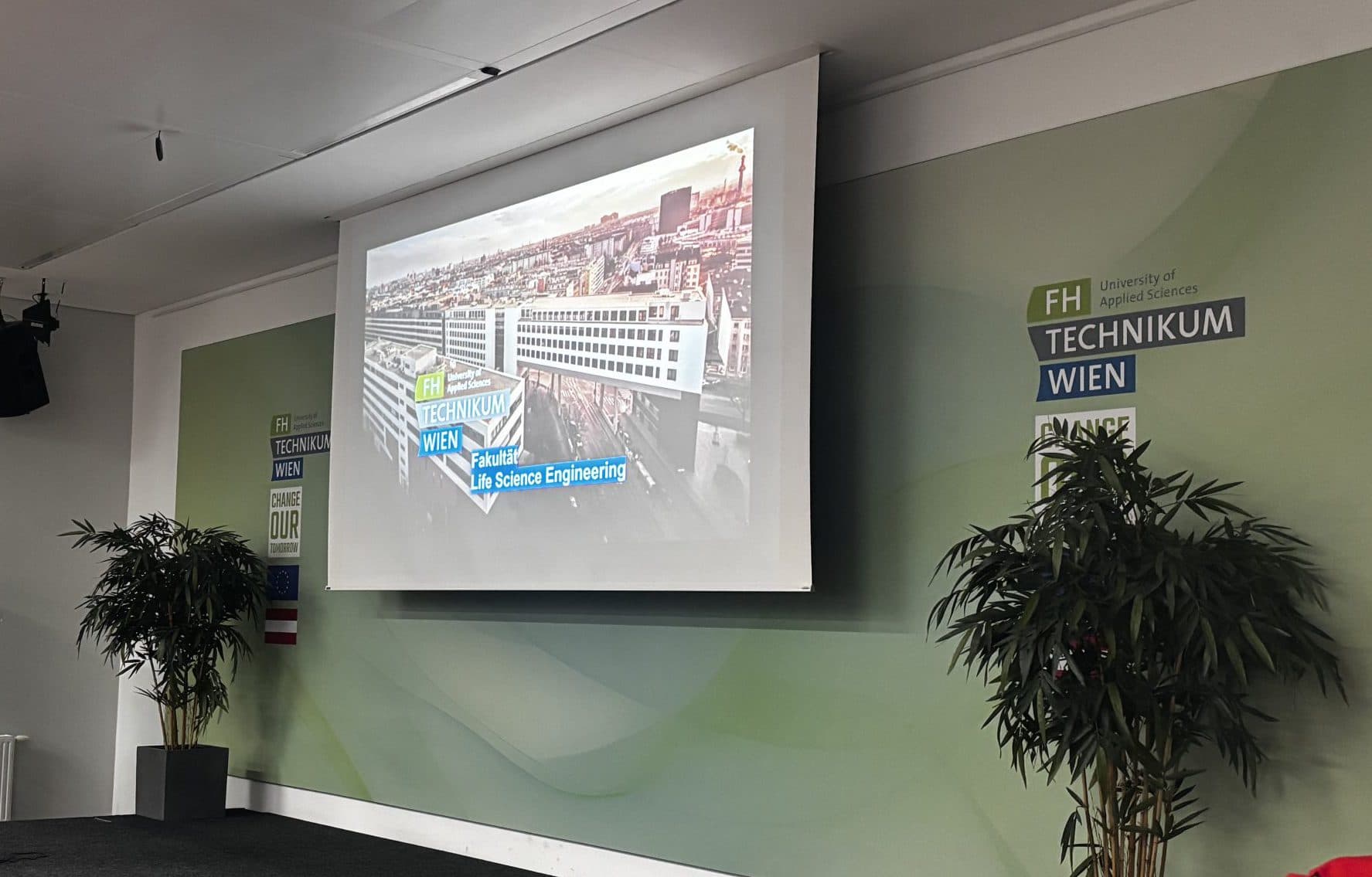 Ein moderner Konferenzraum mit einer Bühne, zwei Topfpflanzen, einem Podium und einer Projektionsfläche, auf der ein Bild einer Stadt mit der Aufschrift Science Engineering zu sehen ist. Das Logo der FH Technikum Wien ist an der Wand angebracht.