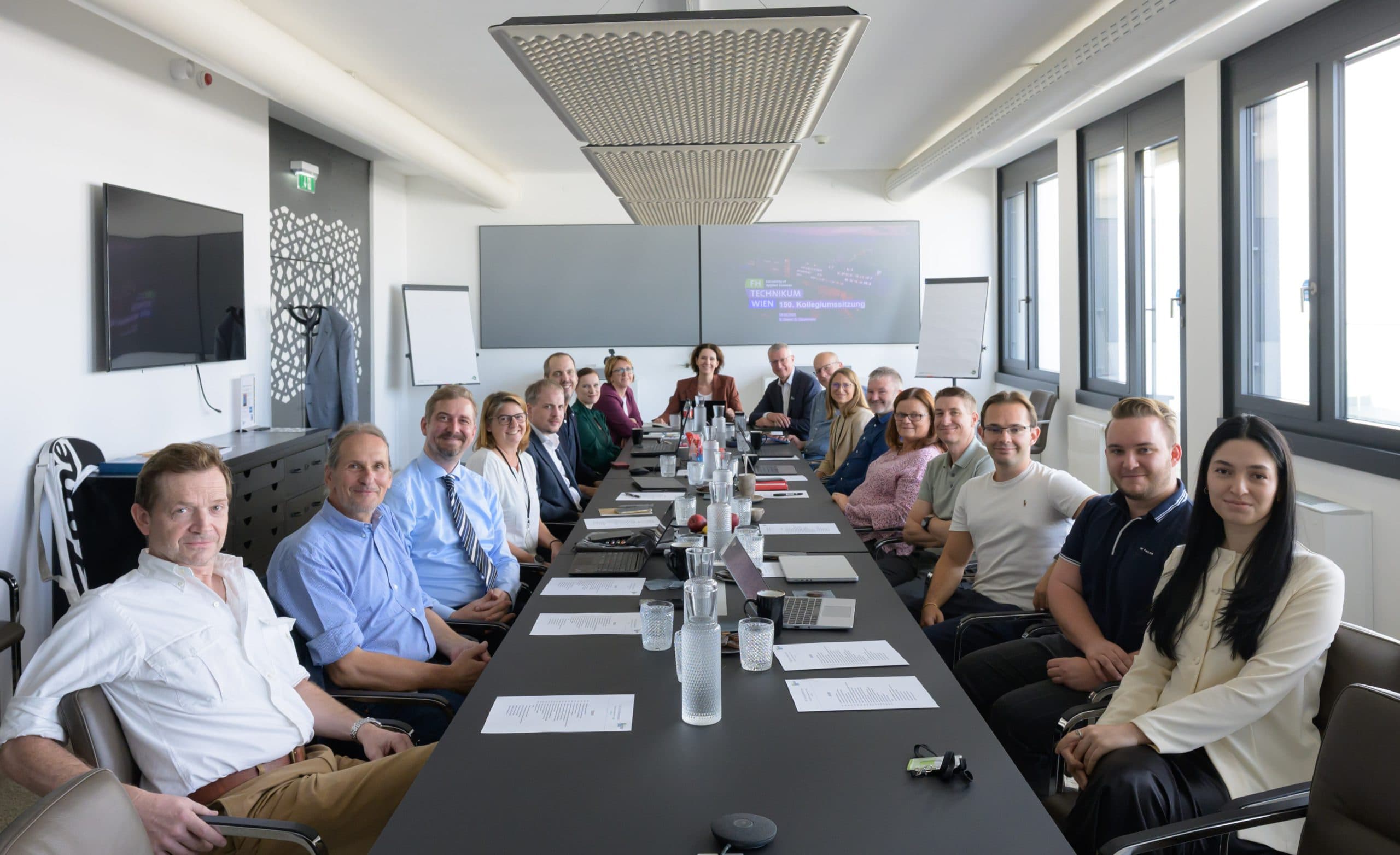 Eine Gruppe von Personen sitzt um einen langen Konferenztisch in einem modernen Besprechungsraum, schaut in die Kamera und lächelt. Auf dem Tisch liegen Papiere, Laptops und Wassergläser. Große Fenster lassen natürliches Licht herein.