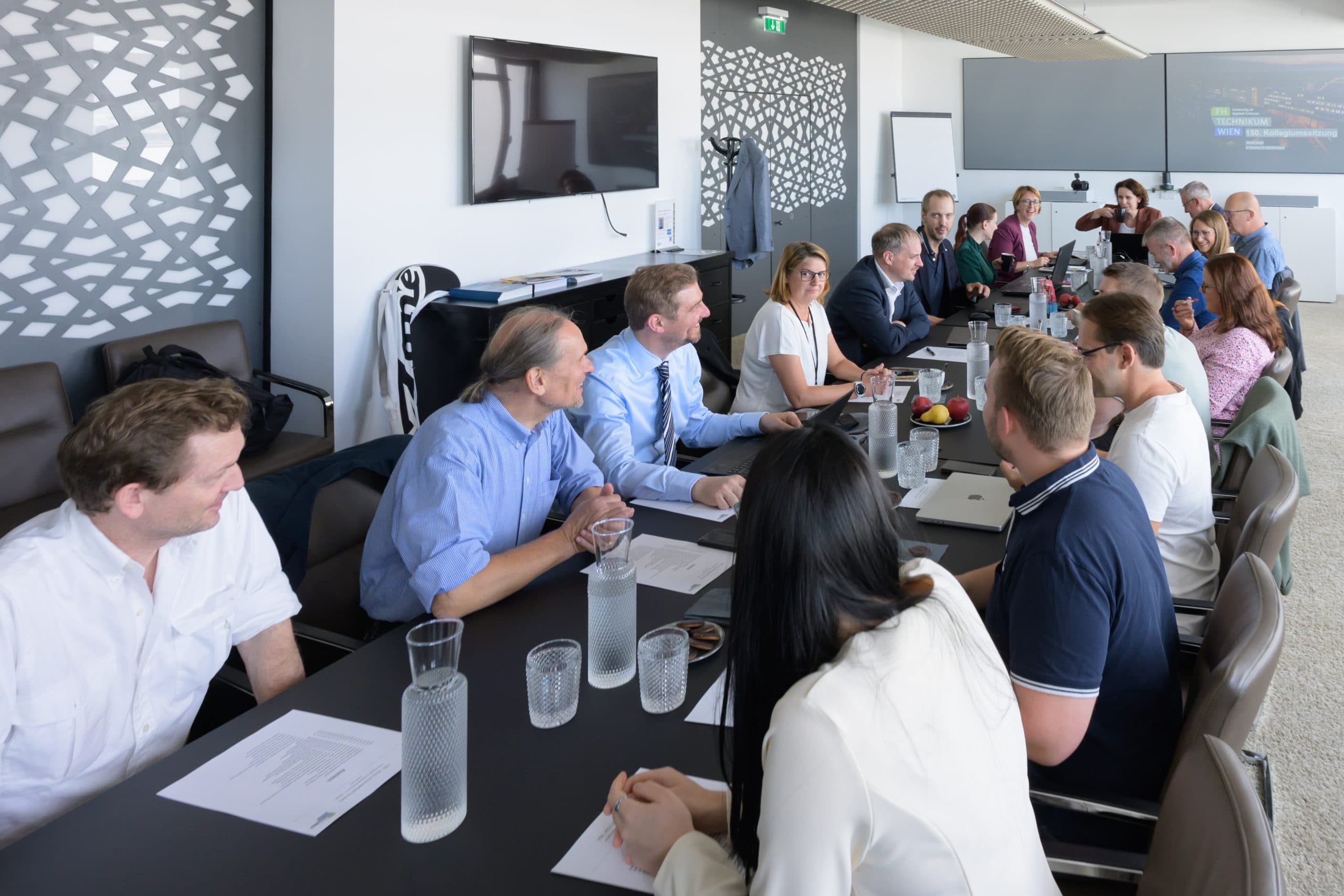 Eine Gruppe von Personen sitzt um einen großen Konferenztisch und führt eine Besprechung durch. Papiere, Wasserkrüge, Gläser und Laptops liegen auf dem Tisch. In der modernen Büroumgebung sind ein Bildschirm und ein Whiteboard zu sehen.