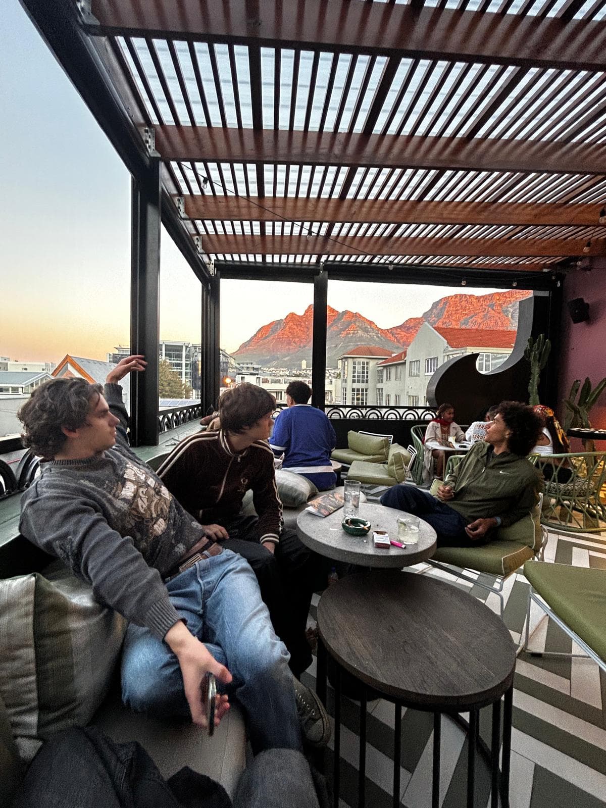 Vier junge Leute entspannen und unterhalten sich in einer Lounge auf dem Dach mit Tischen und gepolsterten Sitzen. Hinter ihnen sind Berge und Gebäude im Licht des Sonnenuntergangs durch ein Pergoladach mit Lamellen sichtbar.