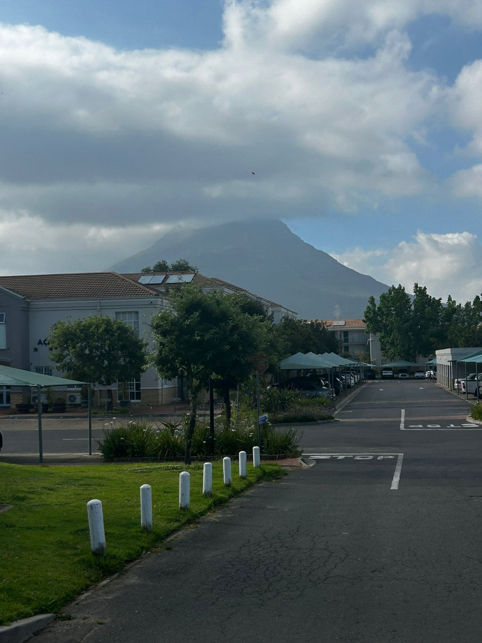 Eine asphaltierte Straße führt an Gebäuden und Bäumen vorbei zu einem entfernten, wolkenverhangenen Berg unter einem teilweise bewölkten Himmel. Weiße Poller säumen die Straße, und an den Seiten sind geparkte Autos zu sehen.