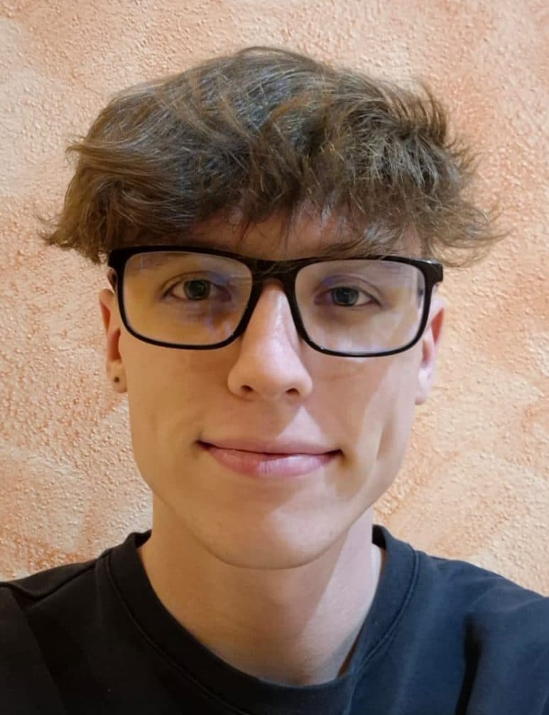 Eine junge Person mit kurzem, gewelltem braunem Haar und Brille lächelt in die Kamera. Sie trägt ein schwarzes Hemd und steht vor einer strukturierten, hellpfirsichfarbenen Wand.