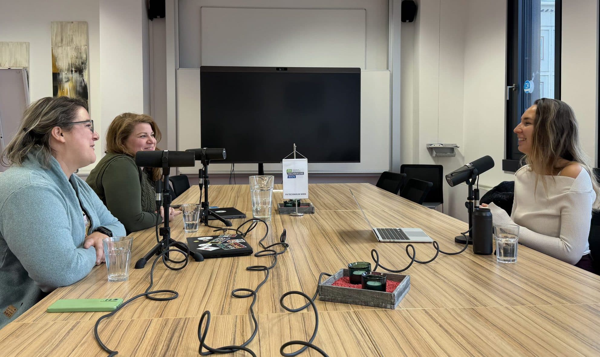 Drei Frauen sitzen mit Mikrofonen um einen Tisch und nehmen in einem modernen, gut beleuchteten Studio einen Podcast auf. Auf dem Tisch stehen Wassergläser, ein Laptop und eine Audioausrüstung. Große Fenster lassen natürliches Licht herein.