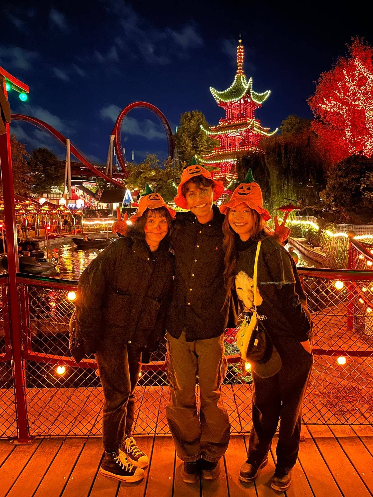 Drei Personen mit Kürbishüten lächeln und posieren zusammen in einem nächtlichen Vergnügungspark, der mit hellen Lichtern, roten Bäumen und einer Pagode geschmückt ist, wobei im Hintergrund eine Achterbahn zu sehen ist.