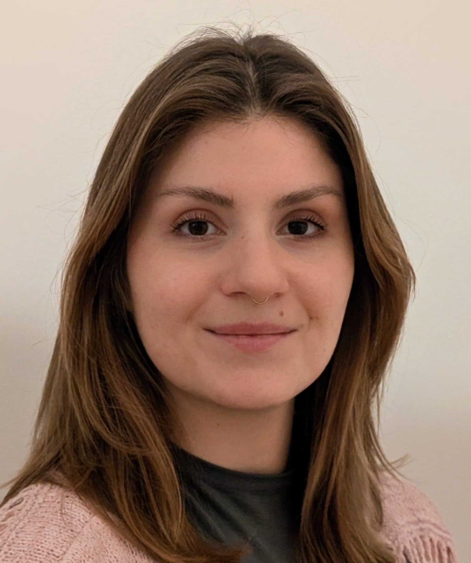 Eine Frau mit glattem braunem Haar, die einen rosafarbenen Strickpullover und darunter ein dunkles Oberteil trägt, lächelt sanft vor einem schlichten hellen Hintergrund.