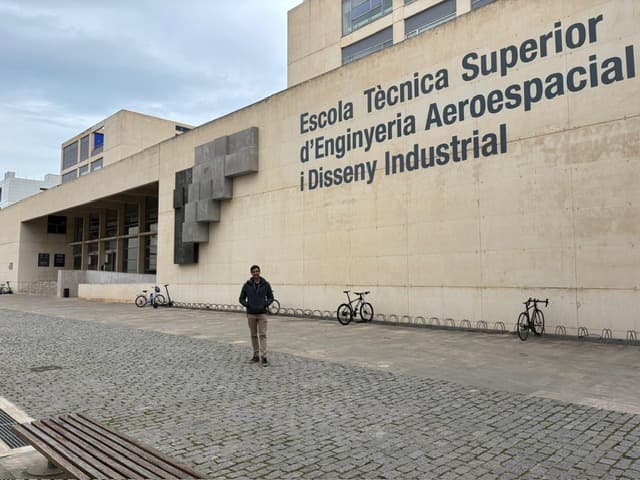Eine Person steht auf einem kopfsteingepflasterten Platz vor einem großen beigen Gebäude mit dem Schild Escola Tècnica Superior dEnginyeria Aeroespacial i Disseny Industrial an der Wand. In der Nähe sind mehrere Fahrräder geparkt.