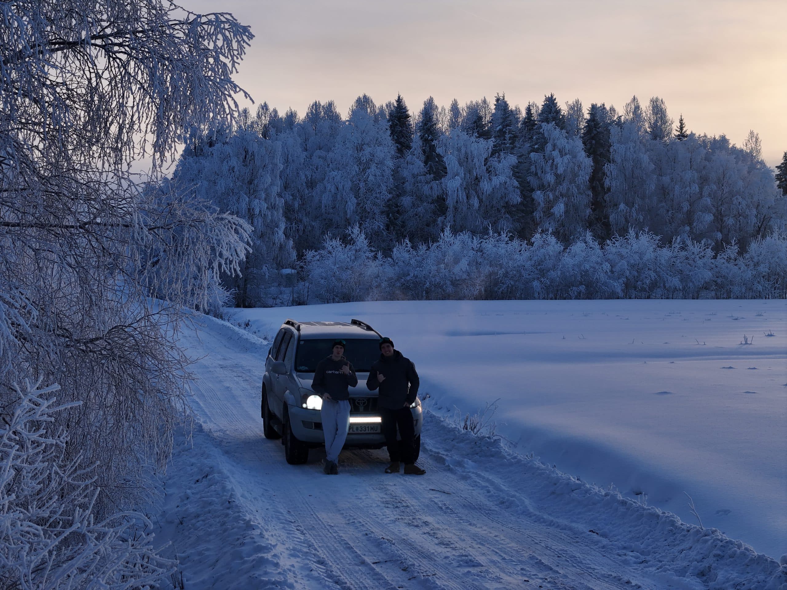 Zwei Personen stehen vor einem Geländewagen, der auf einer verschneiten Straße geparkt ist, umgeben von frostbedeckten Bäumen und einer schneebedeckten Landschaft in der Abend- oder Morgendämmerung. Nebel steigt in der kalten Luft auf, und die Szene hat eine ruhige, winterliche Atmosphäre.