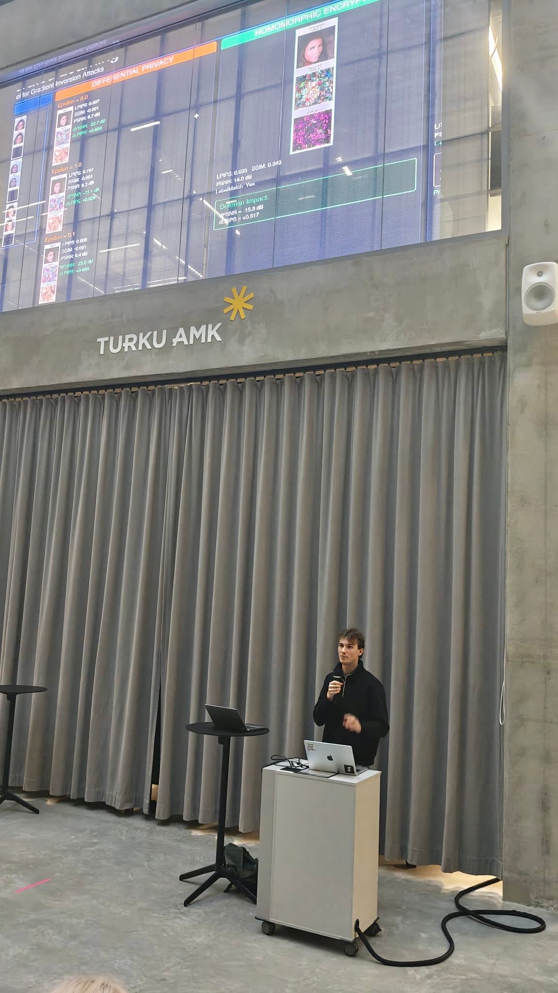 Eine Person steht mit einem Laptop an einem Podium und hält eine Präsentation in einer modernen Halle. Hinter ihr zeigt ein großer Bildschirm bunte Grafiken und Profile. An der Wand steht TURKU AMK über geschlossenen grauen Vorhängen.