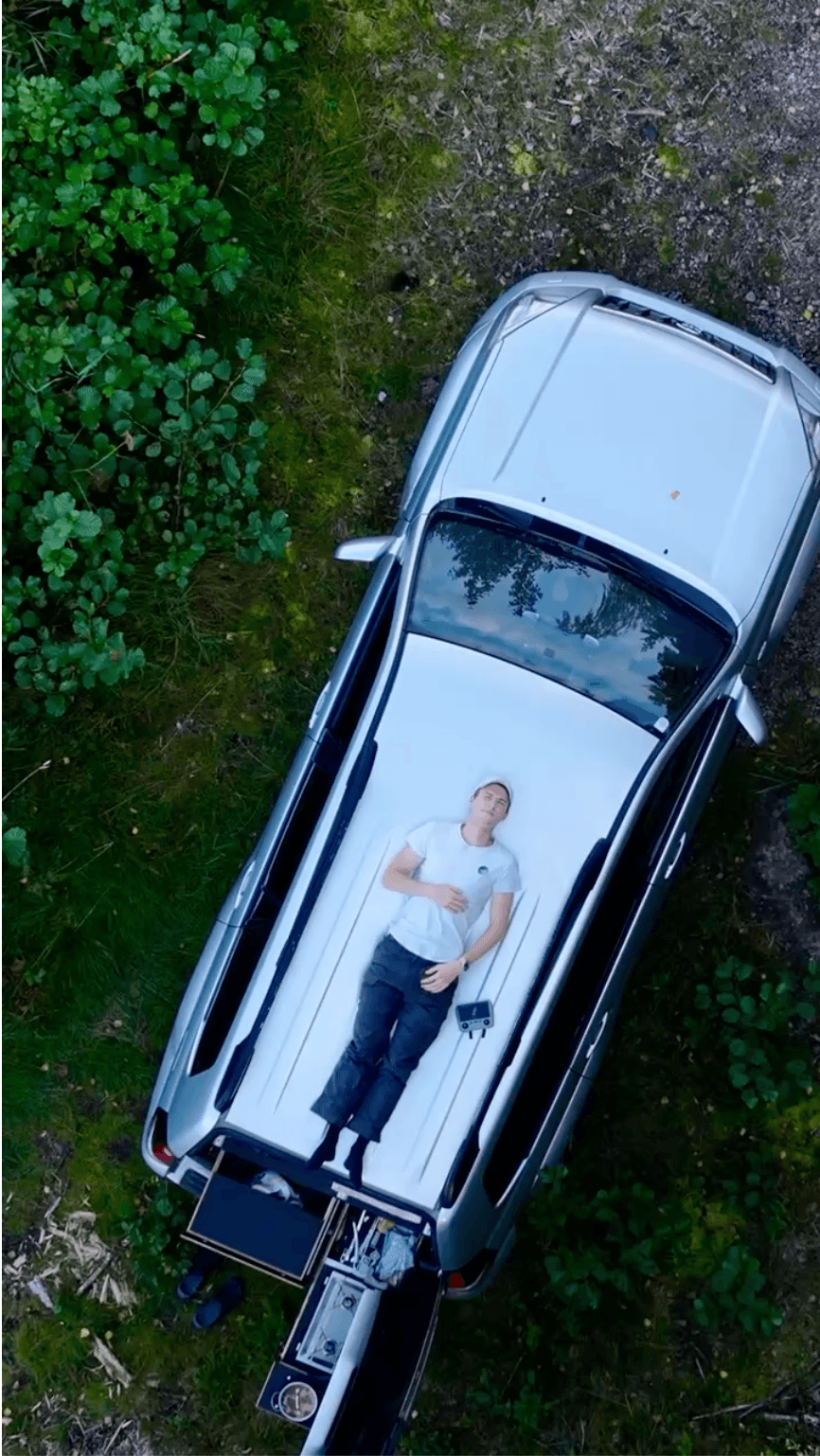 Eine Person liegt auf dem Rücken auf der Ladefläche eines silbernen Pickups, der mit einer weißen Matratze ausgestattet ist, umgeben von grünem Laub und Gras, von oben betrachtet.
