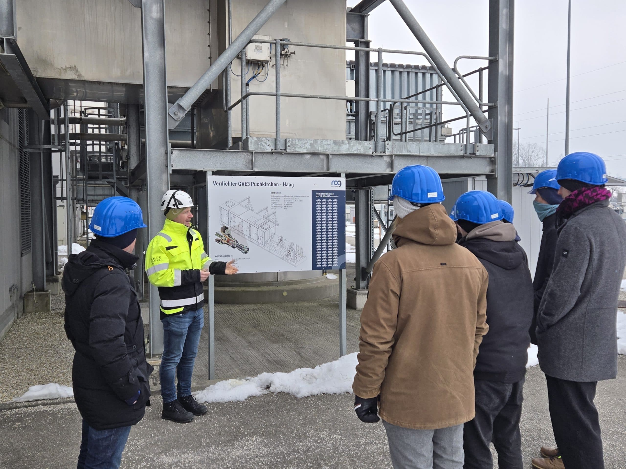 Eine Person mit gelber Sicherheitsjacke und weißem Helm erklärt fünf Personen mit blauen Schutzhelmen und Wintermänteln in einer Industrieanlage im Freien bei Schneefall ein schematisches Diagramm auf einer Tafel.
