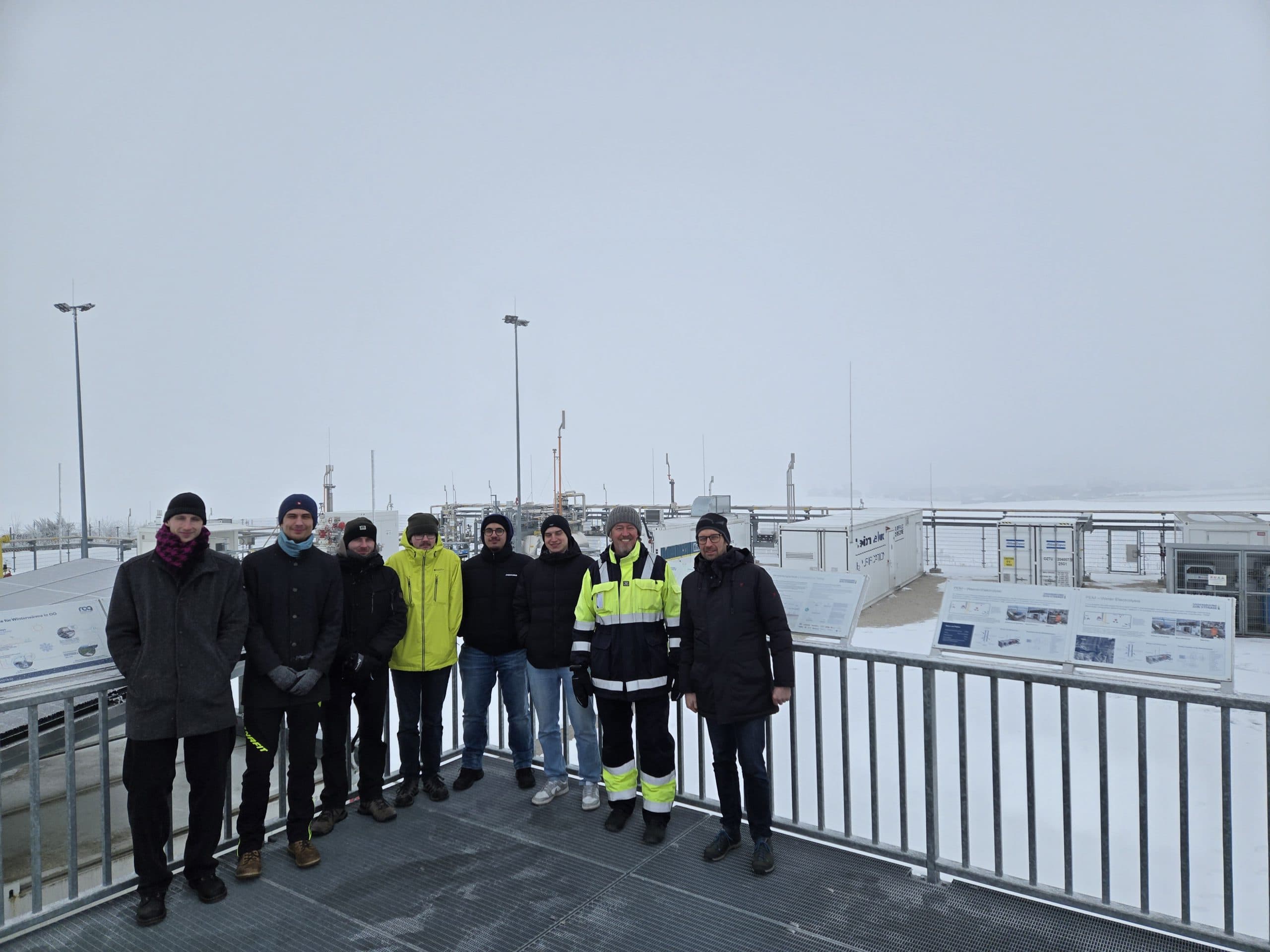 Eine Gruppe von acht Personen in Winterkleidung steht auf einer Metallplattform in einem verschneiten Industriegebiet, im Hintergrund sind Informationstafeln und Geräte zu sehen.