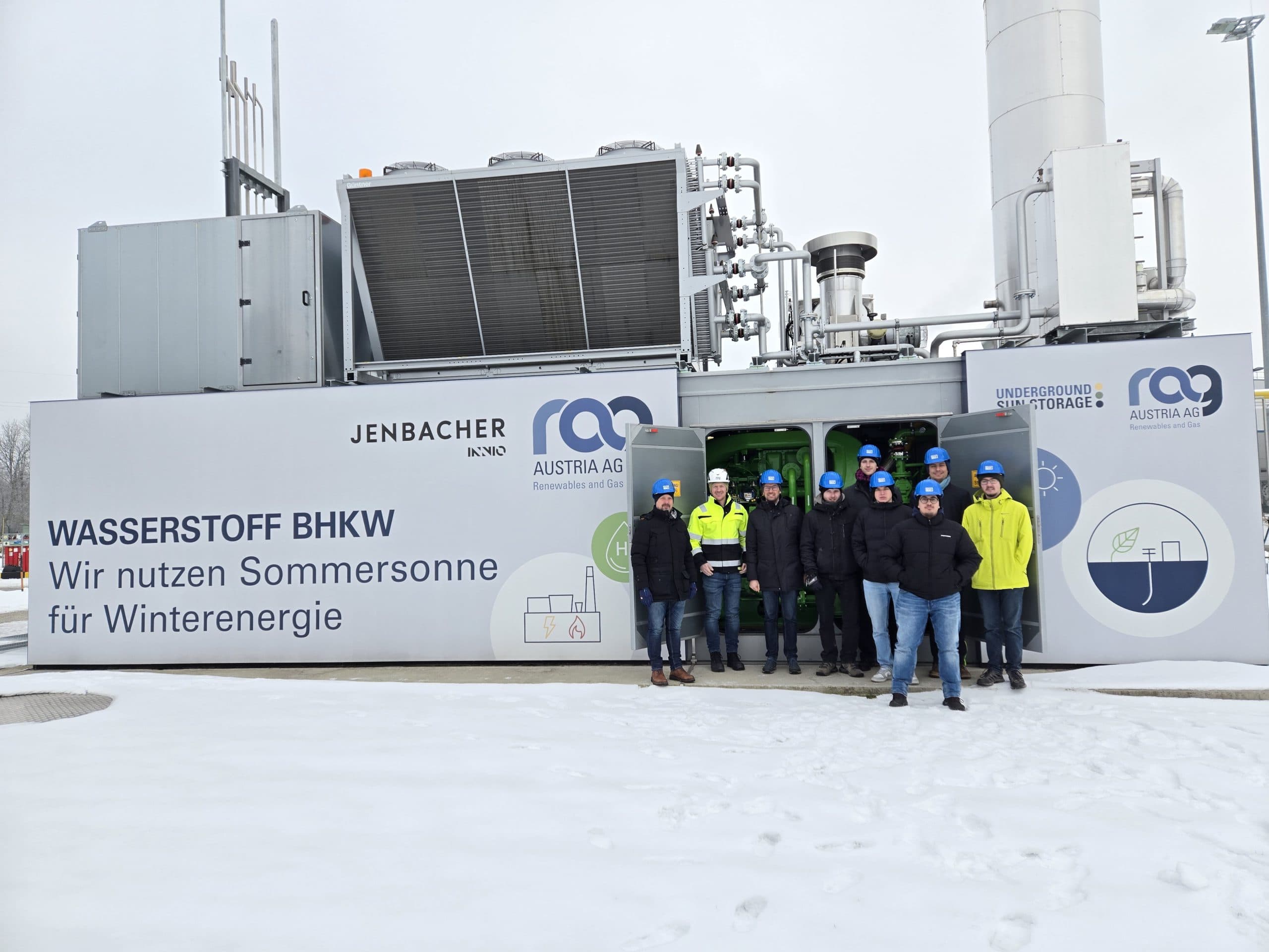 Eine Gruppe von Personen mit Schutzhelmen und Sicherheitsausrüstung steht vor einem Container eines Wasserstoffkraftwerks mit der Aufschrift Wasserstoff BHKW. Der Boden ist mit Schnee bedeckt, und im Hintergrund sind Industrieanlagen zu sehen.